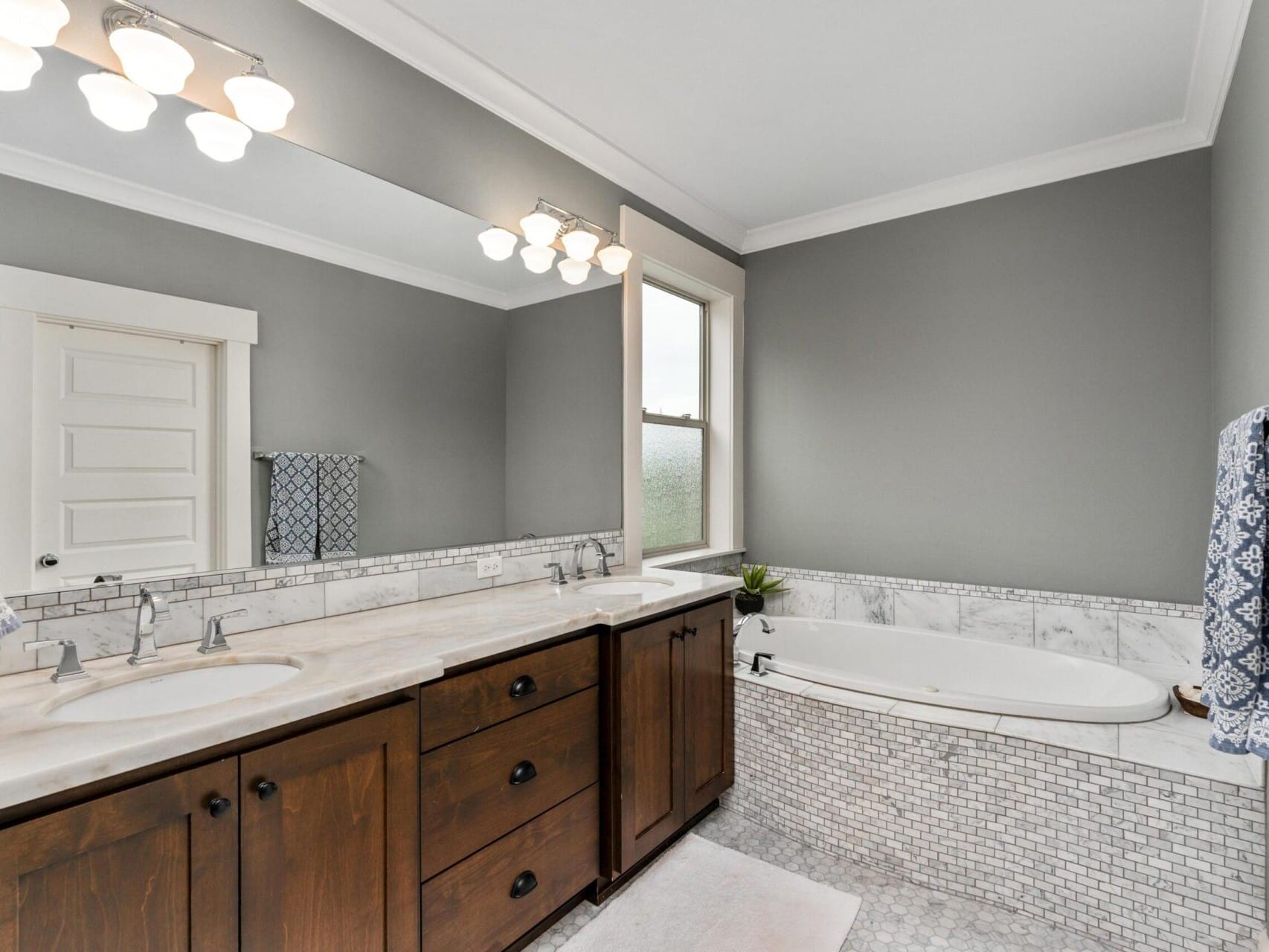 A modern bathroom in a Portland Oregon real estate listing features gray walls, a large mirror, dual sinks with wooden cabinets, and a marble countertop. An oval bathtub is set into a tiled alcove, with frosted glass windows and patterned blue towels adding a touch of decor.