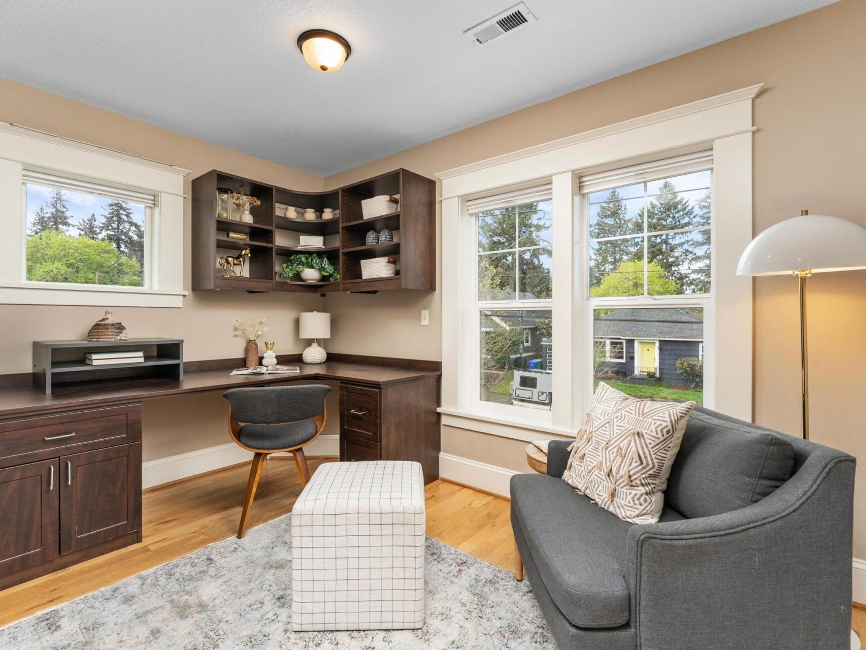 A cozy home office perfect for a Portland Oregon Real Estate enthusiast, featuring a dark wooden desk and shelves, decorative items, and a small chair. A gray sofa with patterned pillows faces a plaid ottoman. Large windows offer views of trees and a yellow house, ideal for any Top Real Estate Agent's inspiration.