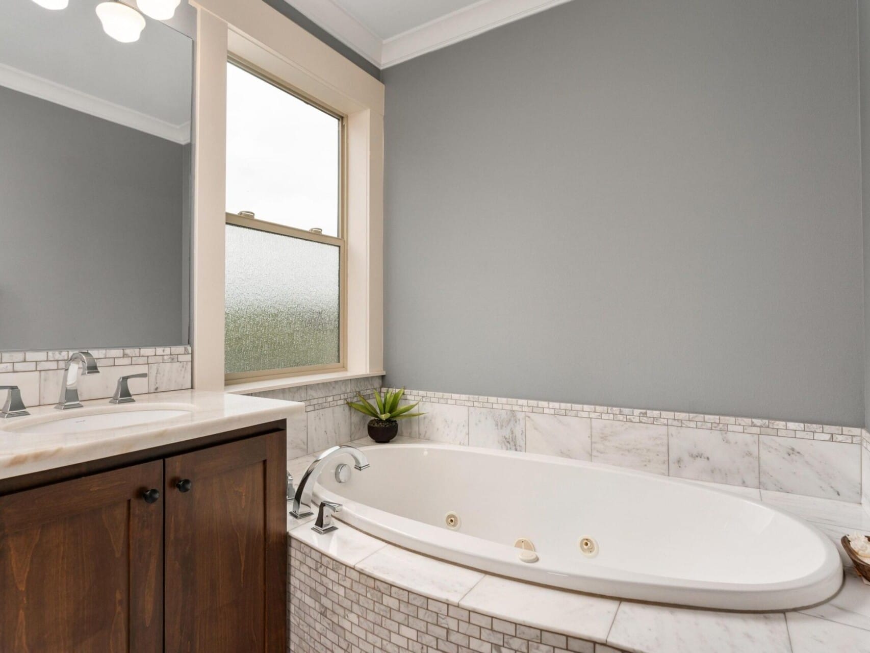 Modern bathroom in Portland Oregon real estate, featuring a large oval bathtub surrounded by marble tiles. A wooden vanity with a sink is on the left, and a small plant sits on the tub ledge. A frosted window allows natural light to fill the space, epitomizing refined elegance curated by a top real estate agent.