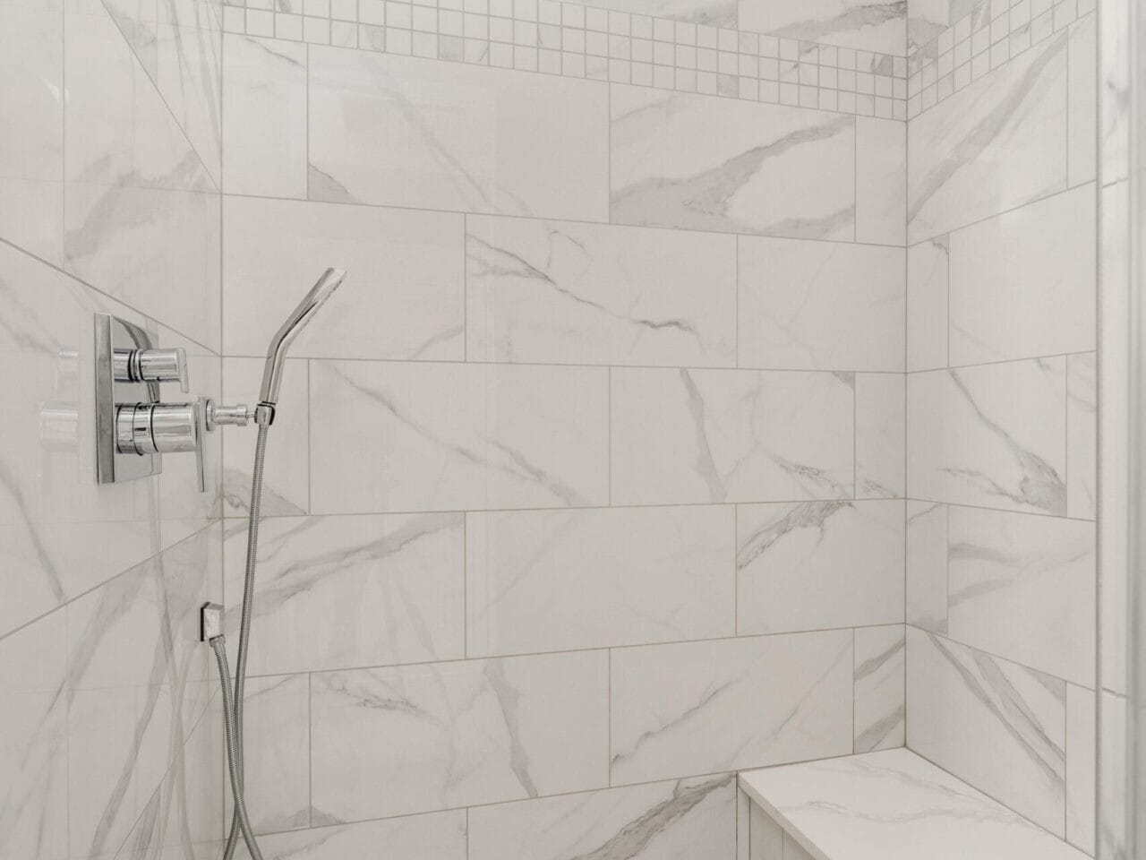 A modern shower in a Portland Oregon real estate gem, featuring large white marble tiles with gray veining. It boasts a square rainfall and handheld showerhead, a built-in bench, and an accent of small square tiles. The floor's elegant white hexagonal tiles complete this luxurious experience.