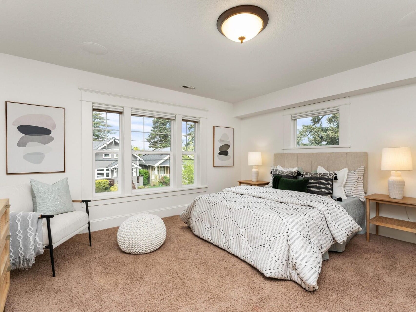 A cozy bedroom in a Portland Oregon Real Estate listing features a large bed adorned with patterned bedding and assorted pillows. Two bedside tables with lamps flank the bed, while a chair and knitted pouf sit near a window showcasing a suburban view. Two abstract art pieces hang on the walls.