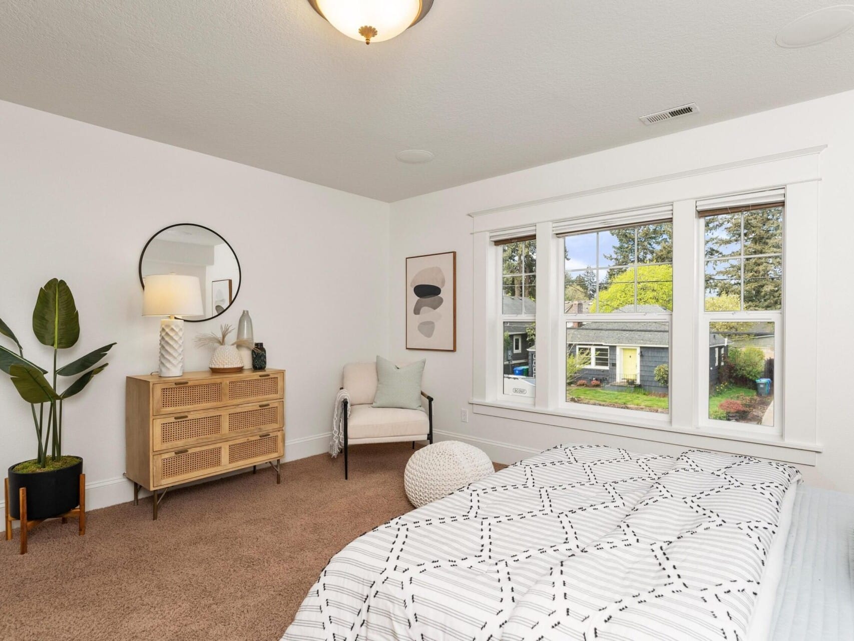 A cozy bedroom in a Portland Oregon real estate gem, featuring a bed with patterned bedding, a wooden dresser, and a round mirror. Enjoy garden views through the large window. Decor includes abstract art and a lamp for added charm, perfect for anyone seeking guidance from a top real estate agent.