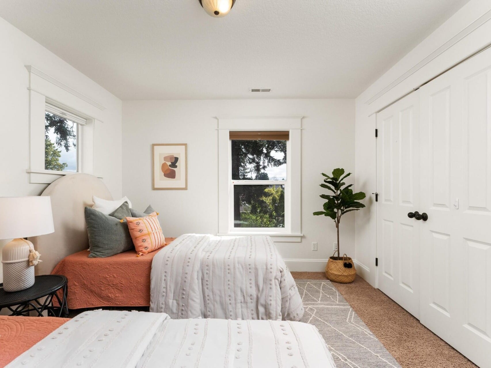 A bright bedroom, perfect for Portland Oregon real estate listings, features two twin beds with white and orange bedding. A round side table with a lamp sits between them. Natural light floods through a large window, while a potted plant adorns the space near the double door closet.