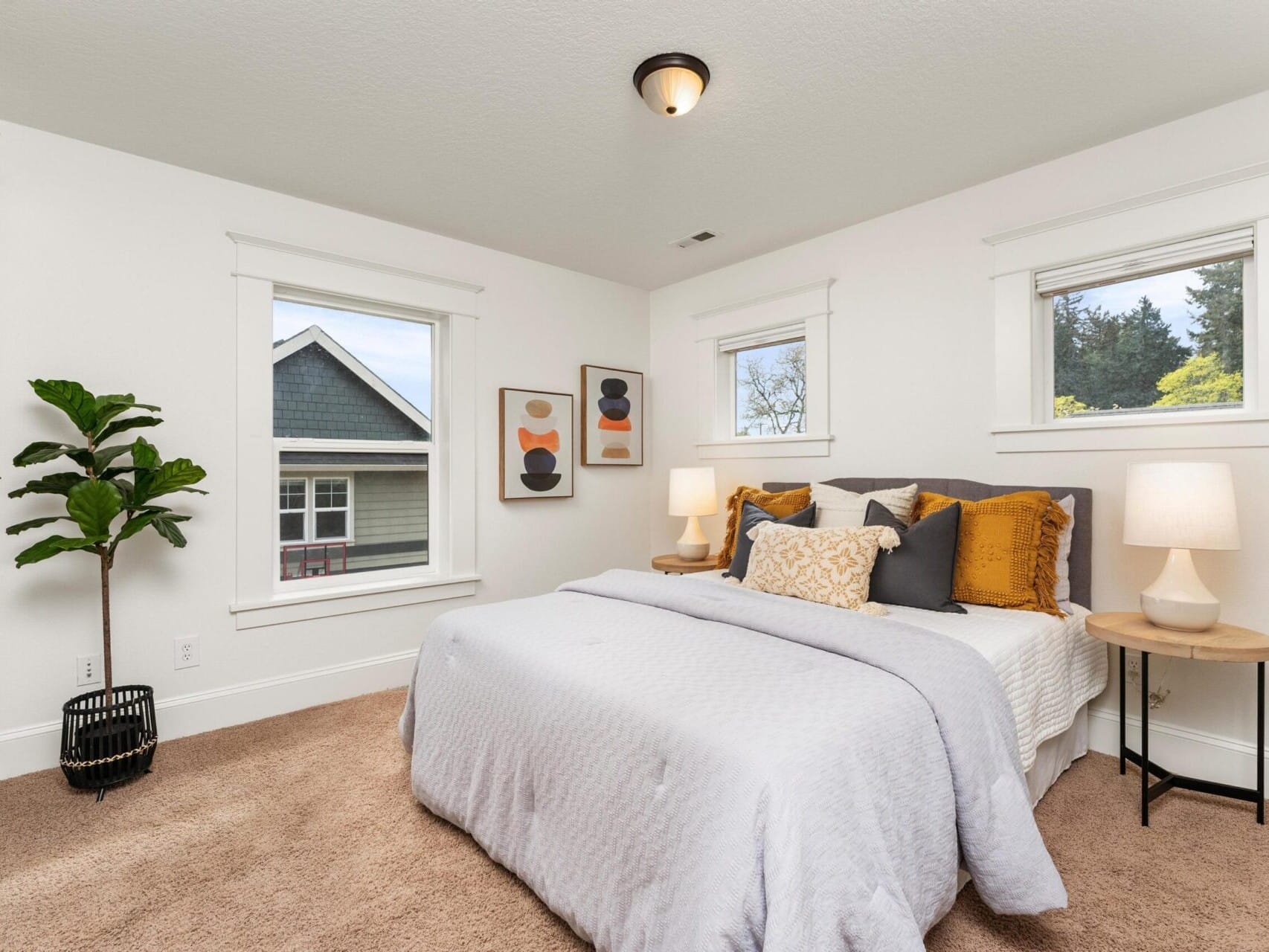 A cozy bedroom in a Portland Oregon real estate listing features a neatly made bed with white and mustard bedding, flanked by wooden nightstands with lamps. A potted plant sits in the corner, and abstract art hangs on the walls. Two windows offer views of a neighboring house and trees.