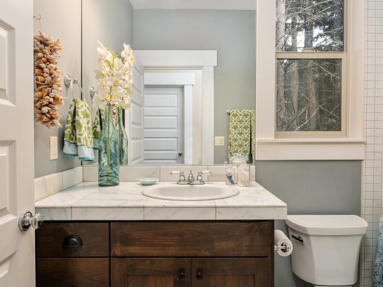 A small bathroom showcases a wooden vanity with a marble countertop, sink, and large wall mirror. Above is a three-bulb light fixture. Decorative plants add charm to the white tiled floor, while a window brightens the space—ideal for anyone exploring Portland Oregon real estate.