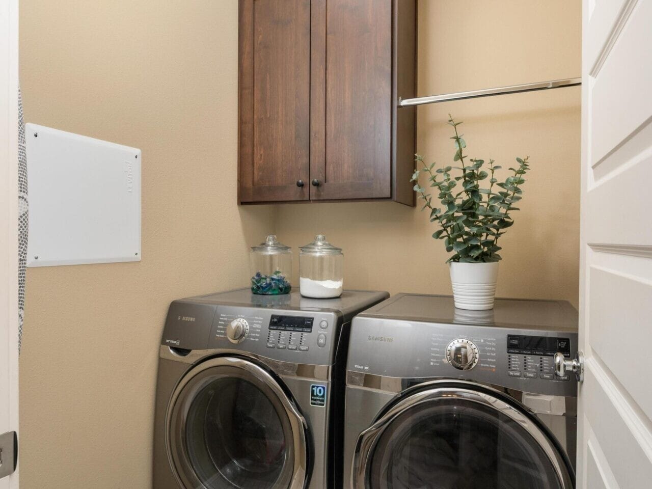 A laundry room in Portland, Oregon real estate showcases a front-loading washer and dryer set on a patterned blue and white tile floor. Above the machines are a wooden cabinet and a shelf with decorative jars and a potted plant. Walls are painted beige, completing this inviting space.