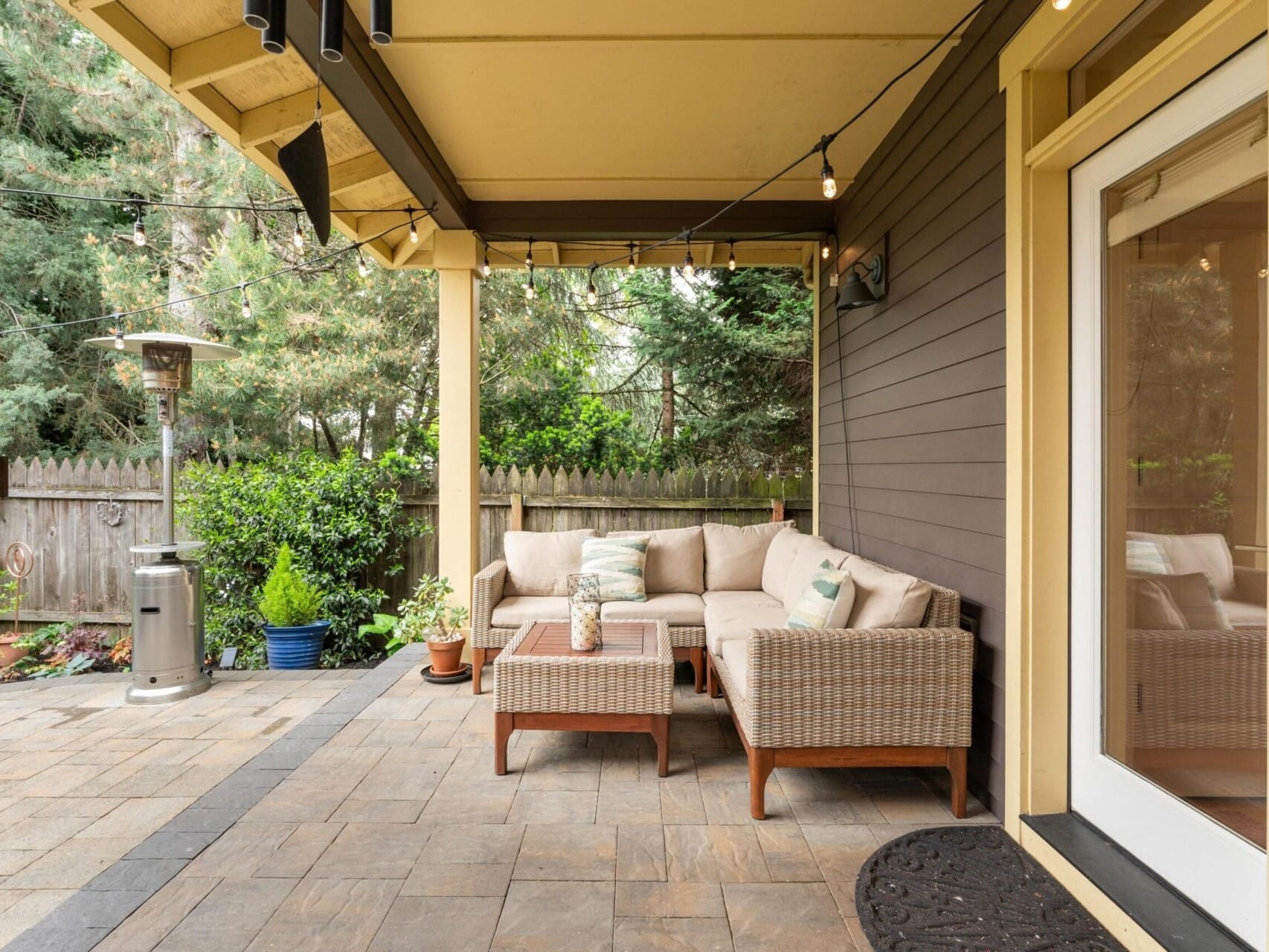 A cozy outdoor patio with a wicker sectional sofa and cushions sits under a wooden pergola. String lights hang above, adding warmth, perfect for a Portland Oregon Real Estate gem. A patio heater stands nearby on the stone floor, while lush green trees and plants border the fenced yard.