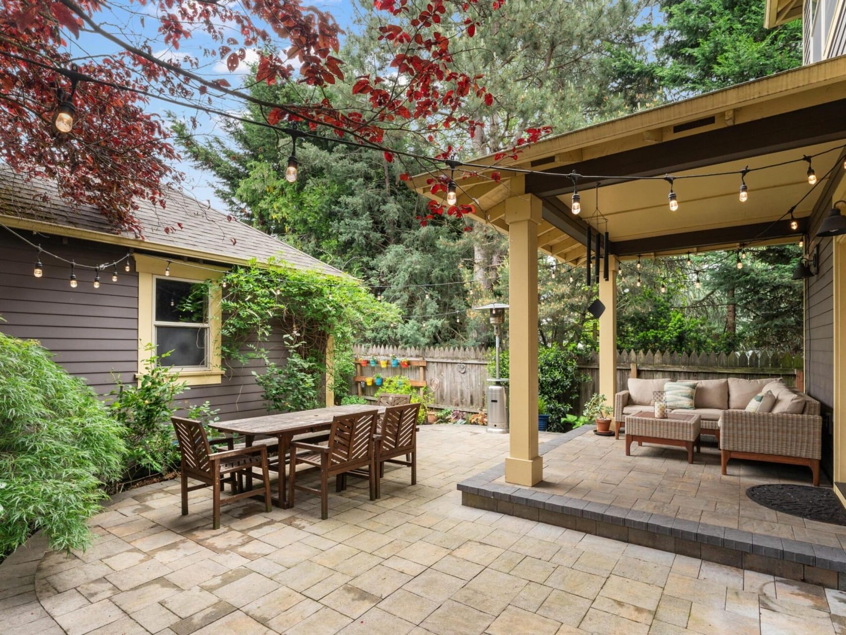 A cozy backyard patio in a Portland Oregon real estate gem, featuring string lights and lush greenery. There's a wooden dining table on the left and a cushioned seating area under a wooden pergola on the right. A shed and a wooden fence elegantly surround this serene space.