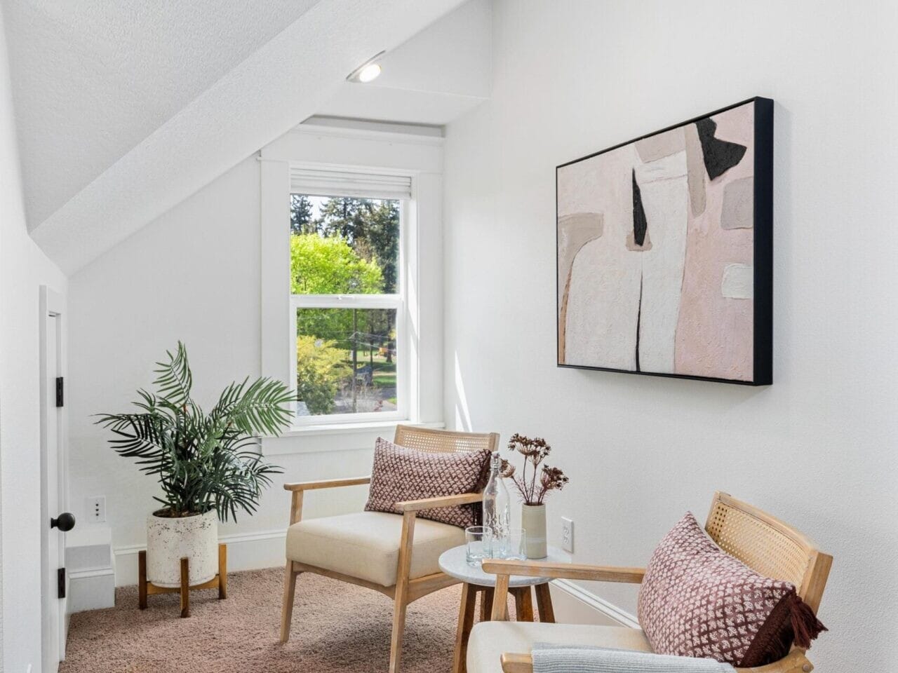 A cozy nook perfect for unwinding, featuring two wooden armchairs with pillows and throw blankets, facing a small round table. A potted plant adds a touch of nature beside them, while an abstract painting graces the wall. Enjoy tranquil views of greenery through the window in this Portland Oregon real estate gem.