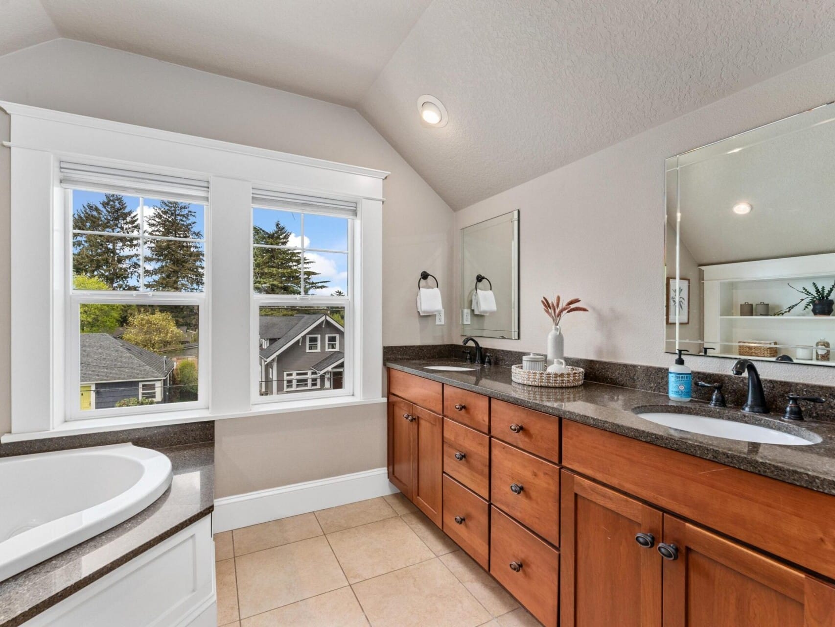 A bright and spacious bathroom with a large window offers picturesque views of trees and houses in Portland. Featuring a double sink vanity with wooden cabinets and a corner bathtub, this gem is just one of the elegant touches highlighted by your top Portland Oregon real estate expert.