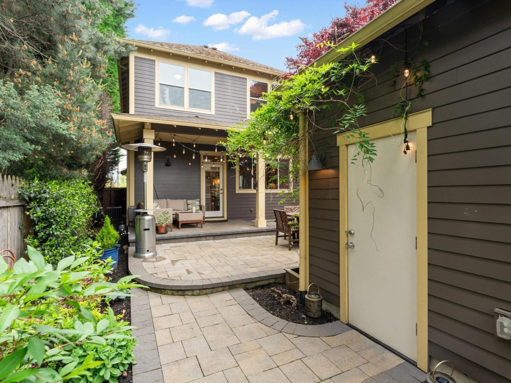 A cozy backyard features a patio with string lights, a seating area, and a view of a two-story house with a covered porch. Surrounded by lush greenery, it creates a tranquil atmosphere—a perfect find in Portland Oregon real estate, ideal for those seeking peace and beauty at home.