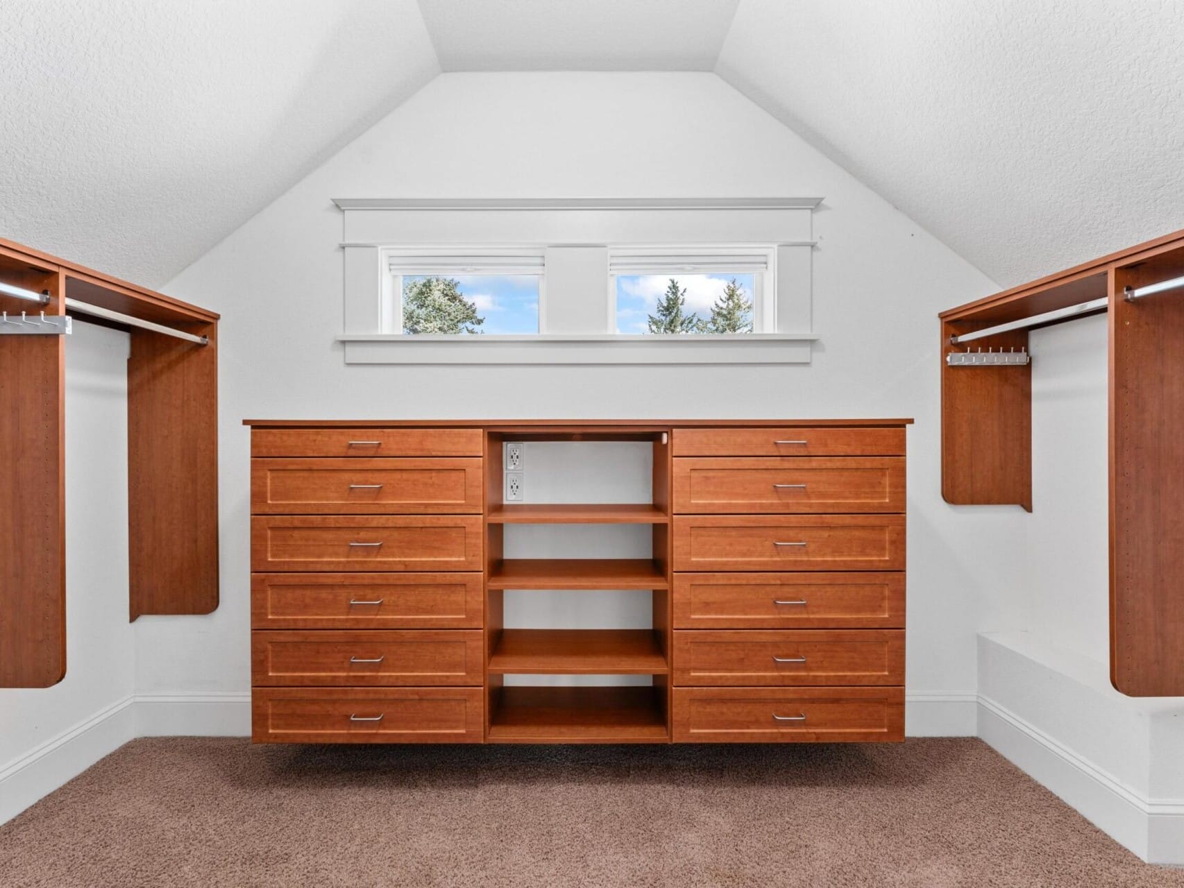 A spacious walk-in closet with an angled ceiling, crafted for those exploring Portland Oregon real estate. It features built-in wooden cabinets with multiple drawers in the center, surrounded by hanging rods on each side. Two rectangular windows above the cabinets let in natural light.