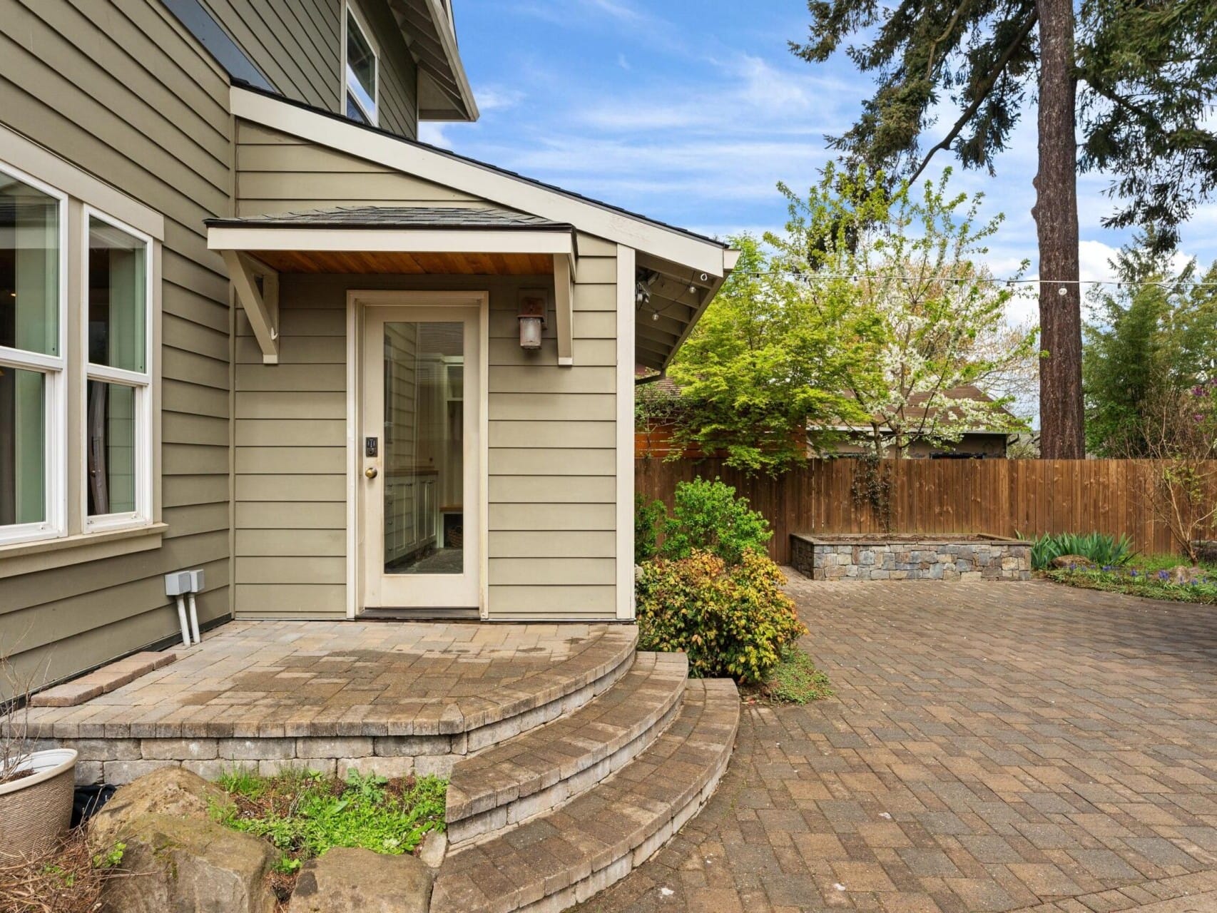 Exterior view of a Portland real estate gem, showcasing green siding and a glass door. A small stone patio with steps complements the scene, while a wooden fence borders the yard with shrubs and a large pine tree. The partly cloudy sky enhances this quintessential Portland Oregon home.