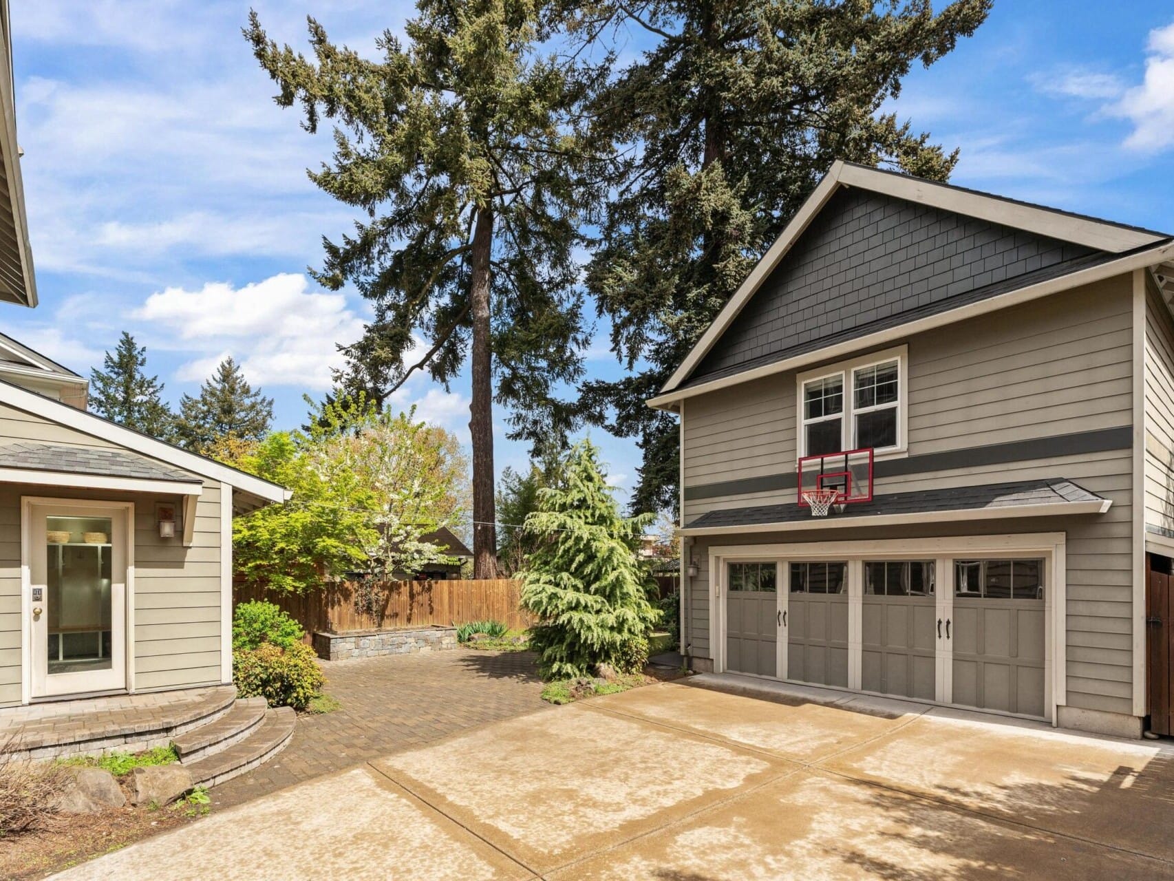 A spacious driveway leads to a two-story gray house with a basketball hoop above the garage doors. Tall evergreen trees and a wooden fence surround the property, set against Portland, Oregon's clear blue skies—a gem in Portland real estate.