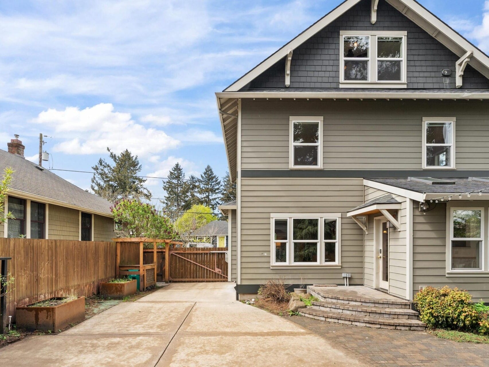 A gray two-story house with white trim and multiple windows, nestled in the desirable Portland Oregon real estate market. It features a small porch with brick steps and a paved driveway leading to a wooden fence. The sky is clear with a few clouds, and trees and shrubs enhance the property’s charm.