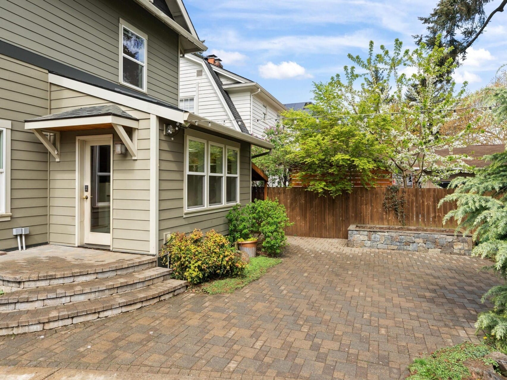 A charming two-story house with green siding awaits in the heart of Portland Oregon real estate. It boasts a small stone patio and paver driveway. Lush greenery, including tall trees and bushes, surrounds the area. A wooden fence encloses the backyard, complete with a stone planter near the fence.