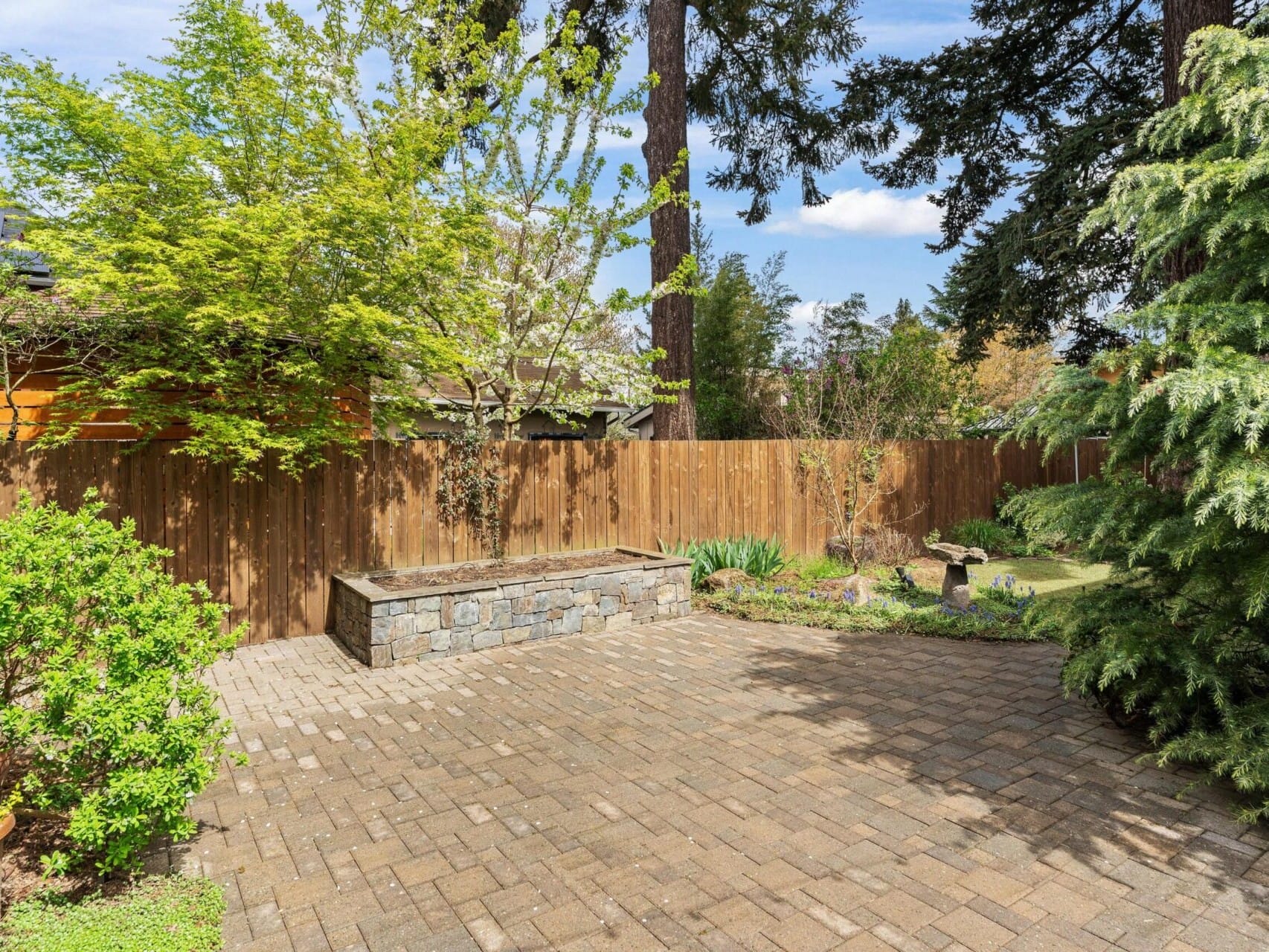 A sunny backyard in Portland, Oregon real estate features a stone-lined planter box surrounded by tall trees and a wooden fence. The brick-paved ground and lush greenery create a serene outdoor space, perfect for anyone seeking tranquility with the help of a top real estate agent.