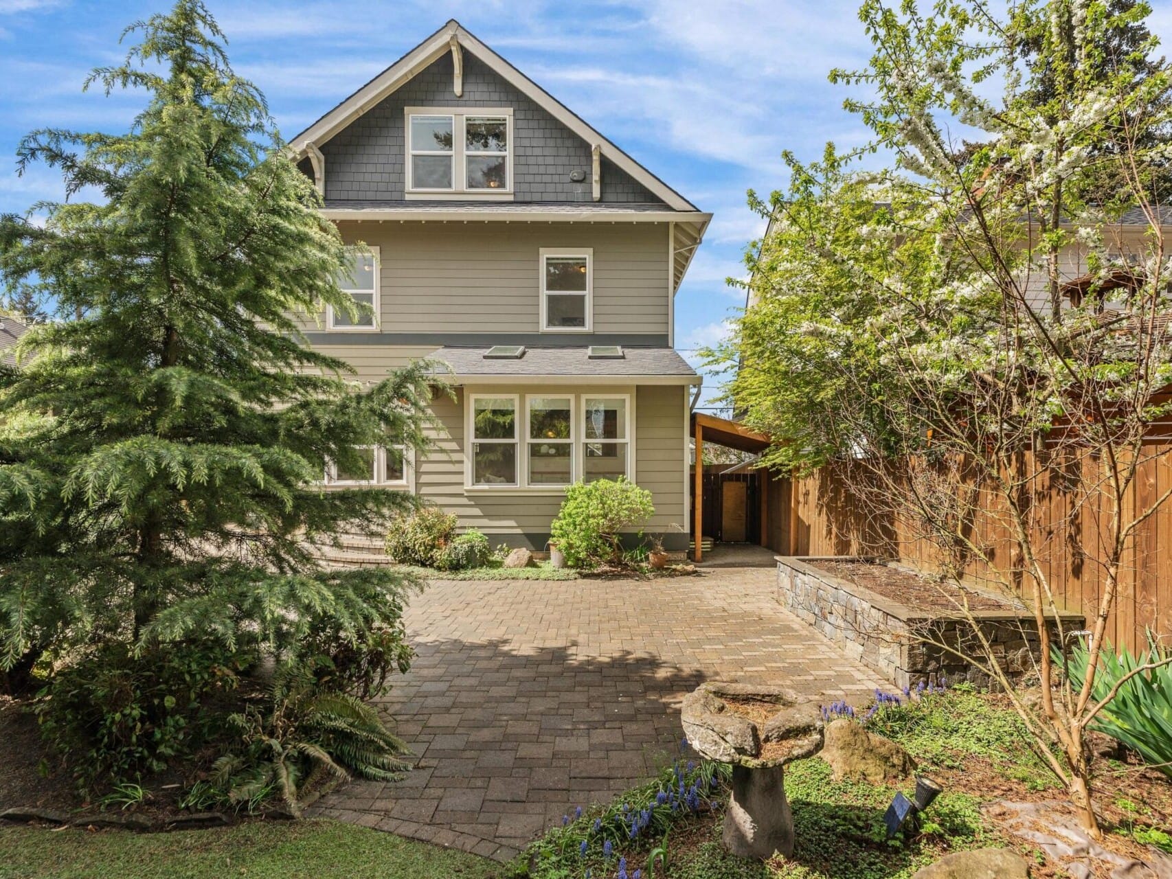 A two-story house with gray siding and a peaked roof is nestled in a landscaped garden, showcasing the best of Portland Oregon real estate. A paved path leads to the entrance, flanked by a tall evergreen tree and flowering plants. A wooden fence is visible on the right side.