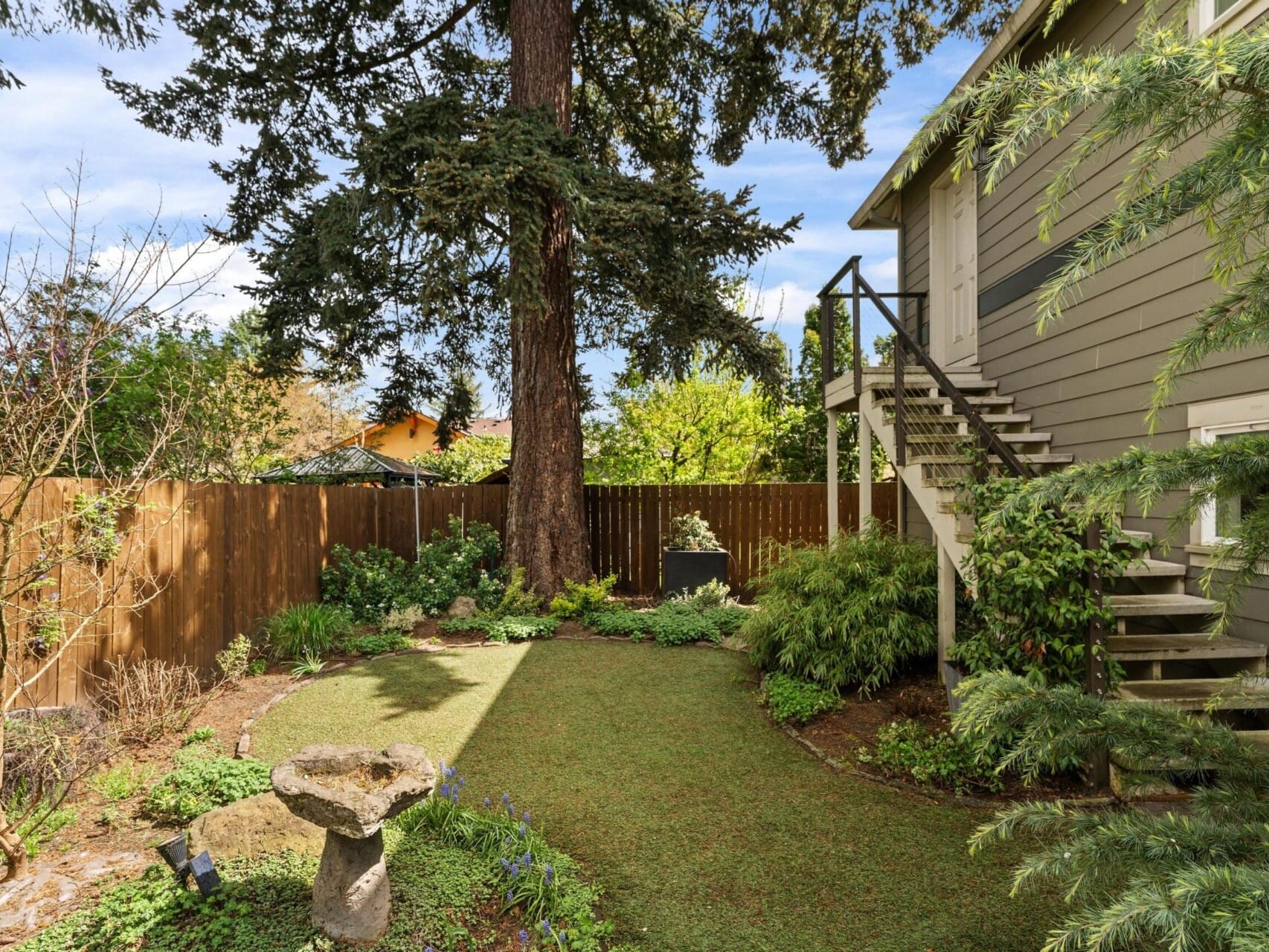 A backyard with a wooden fence, a large tree, various plants, and a birdbath on a patch of green lawn. A set of stairs leads to the second floor of a gray house. Perfectly showcasing the charm found in Portland Oregon real estate, this scene is beautifully illuminated by daylight.