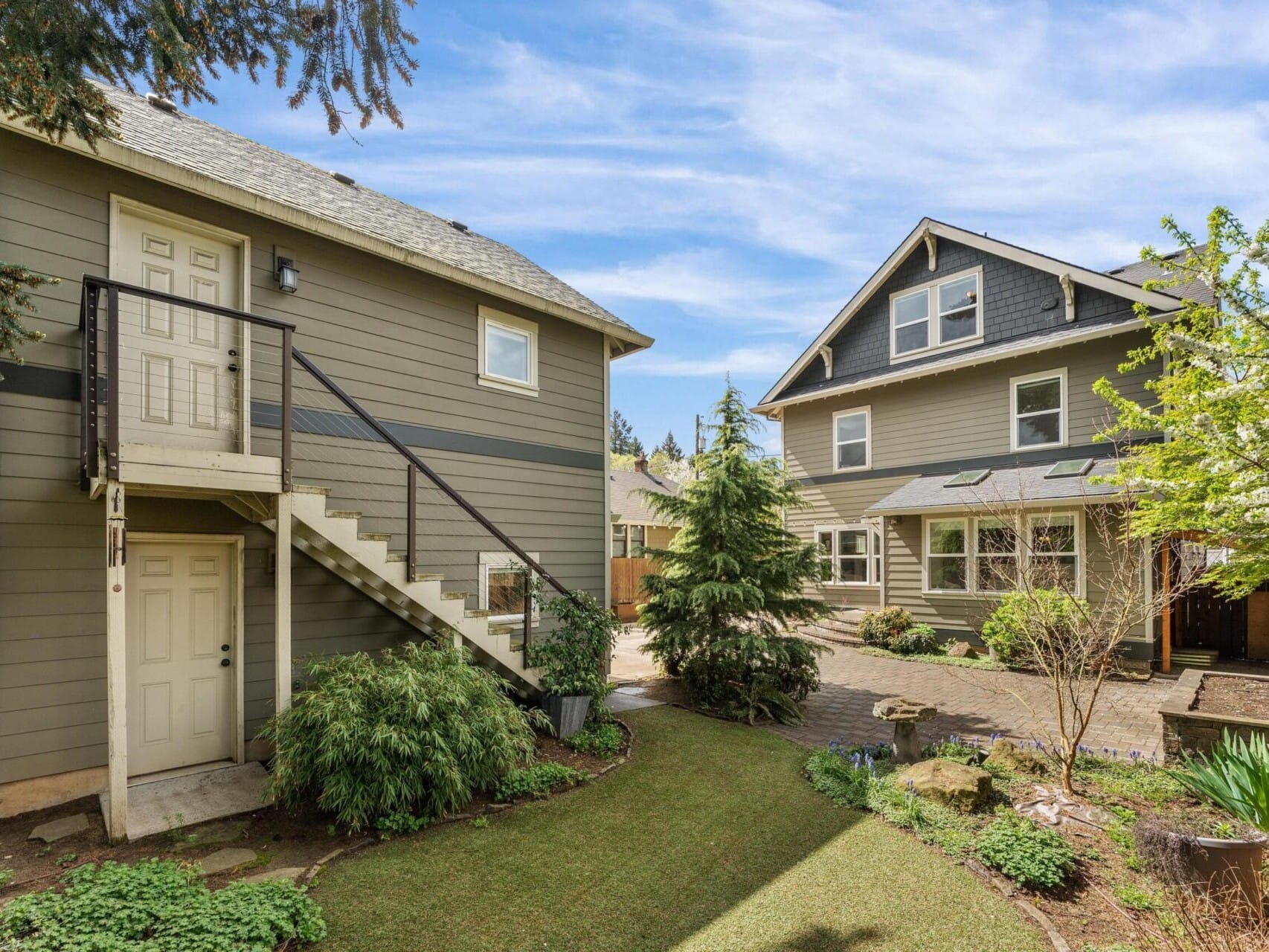 A two-story house with gray siding and white trim showcases its charm, featuring a staircase on the left leading to a door. The well-maintained garden, filled with bushes and trees, is perfect for anyone exploring Portland Oregon real estate under a vibrant blue sky dotted with clouds.