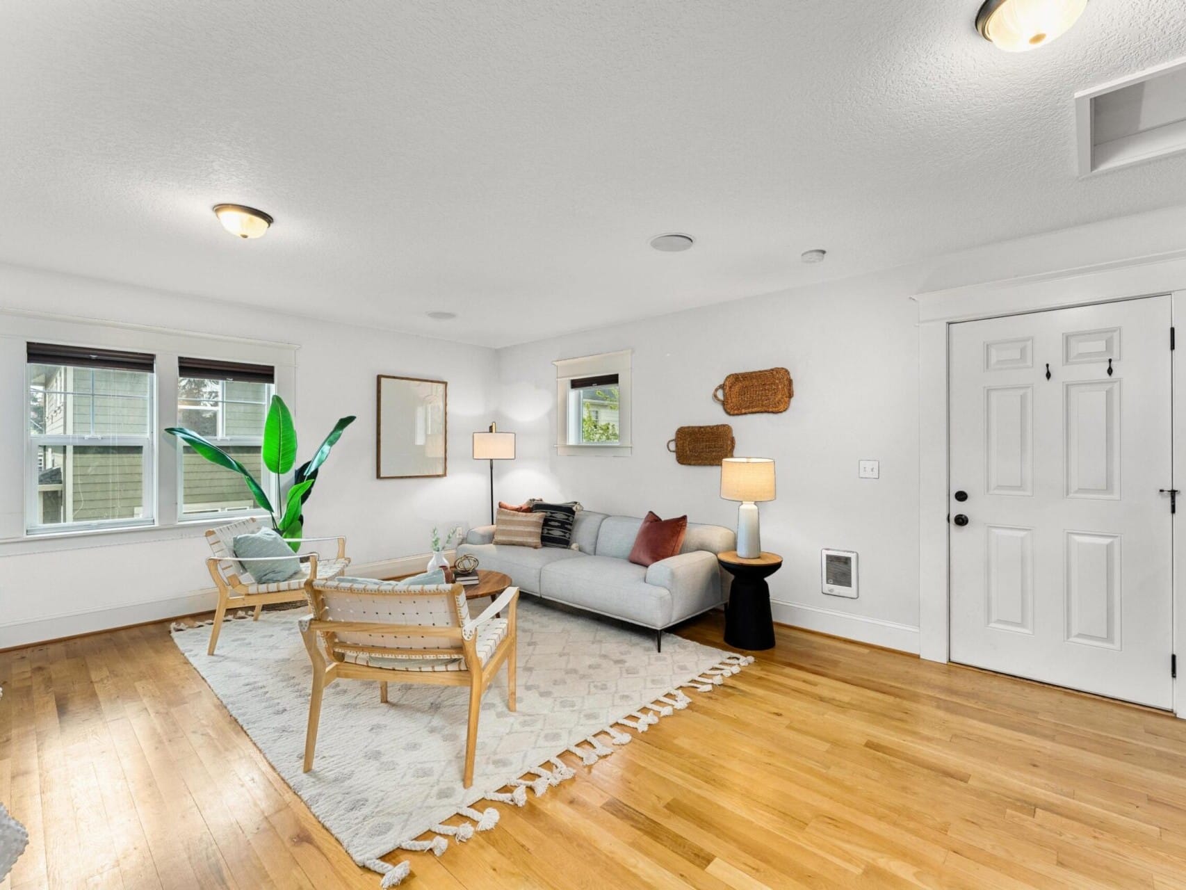 A cozy living room in Portland, Oregon, with wooden flooring features a white sofa, two wooden chairs, and a modern coffee table on a light patterned rug. Natural light fills the space through three windows. This inviting room could be yours with guidance from a top Portland realtor.