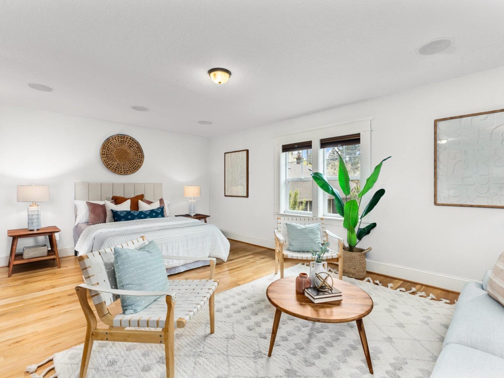 A bright and spacious bedroom with a large bed, patterned pillows, and a woven headboard. Two chairs and a round wooden coffee table grace the foreground. Large windows, a potted plant, and minimalist decor create a calm atmosphere perfect for any discerning Portland realtor's client.