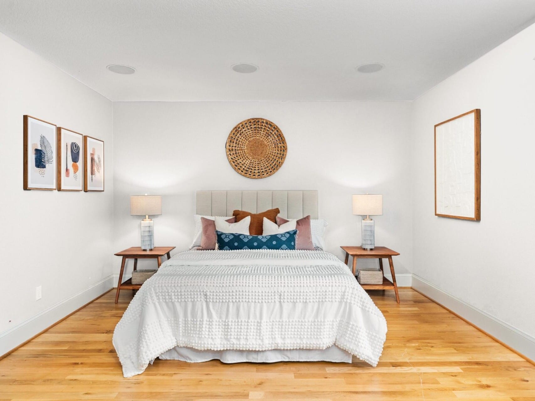 A bright bedroom in Portland Oregon real estate showcases a neatly made bed with white and patterned pillows, flanked by wooden side tables with lamps. Framed artwork and a large woven decor piece adorn the walls, while light wood floors complete this inviting space.