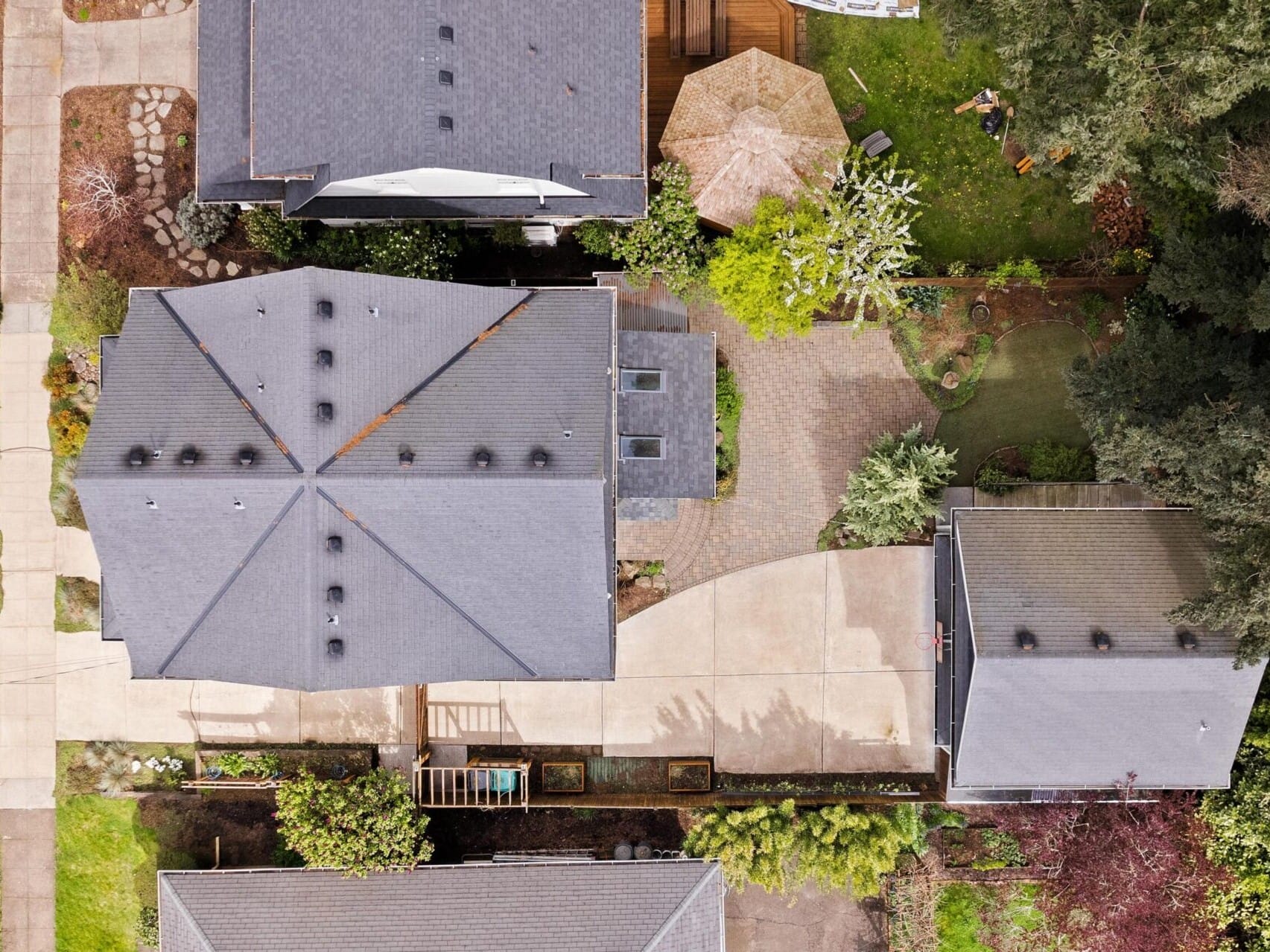 Aerial view of a residential area with three houses. The central house boasts a gray roof, a curved driveway, and a small garden. Surrounded by trees and greenery, a path leads to the entrance—perfect for those exploring Portland Oregon real estate options.