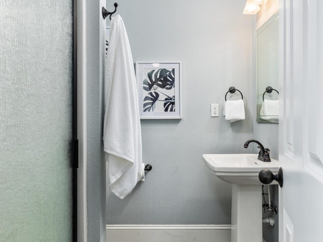 A small bathroom in a Portland Oregon real estate gem features a shower with textured glass, a white pedestal sink, and a framed botanical print. A light fixture above the mirror complements the gray walls and tiled floor in brown and blue tones.