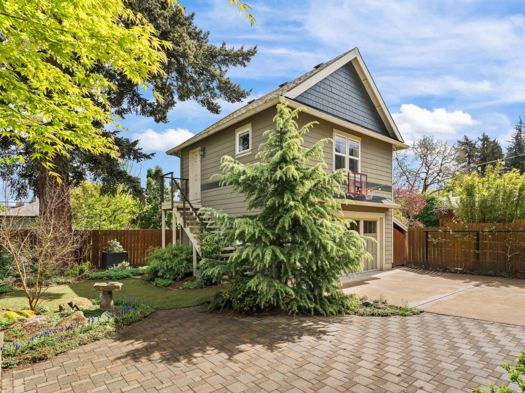 A charming two-story house with gray siding and white trim is nestled in Portland's vibrant real estate scene. The lush garden, brick driveway, and staircase to the second floor add to its allure. Tall trees and a clear blue sky complete this picturesque setting for a top real estate agent.