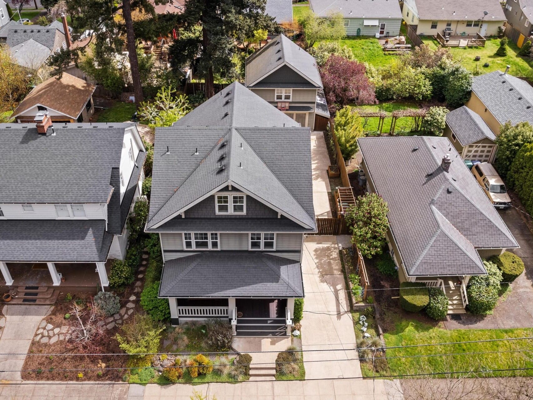 Aerial view of a Portland Oregon neighborhood reveals several houses, with the central one boasting a gray roof and cozy front yard. Surrounded by similar architecture, trees, and gardens, it's a prime example of Portland real estate that any top Realtor would appreciate.