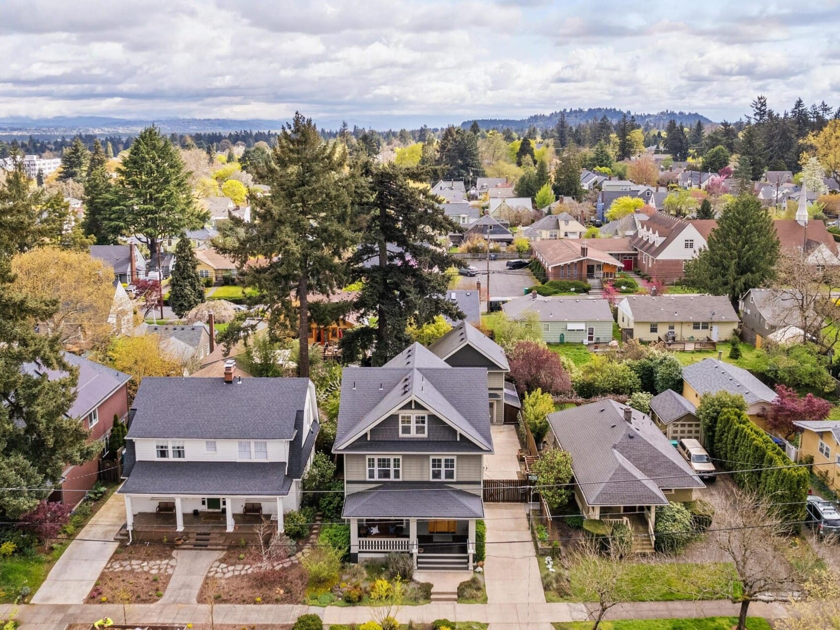 Aerial view of a residential neighborhood in Portland, Oregon, with houses surrounded by trees. The scene showcases greenery and a distant landscape under a partly cloudy sky, reflecting both traditional and modern architecture styles—a perfect setting for any top real estate agent to explore.