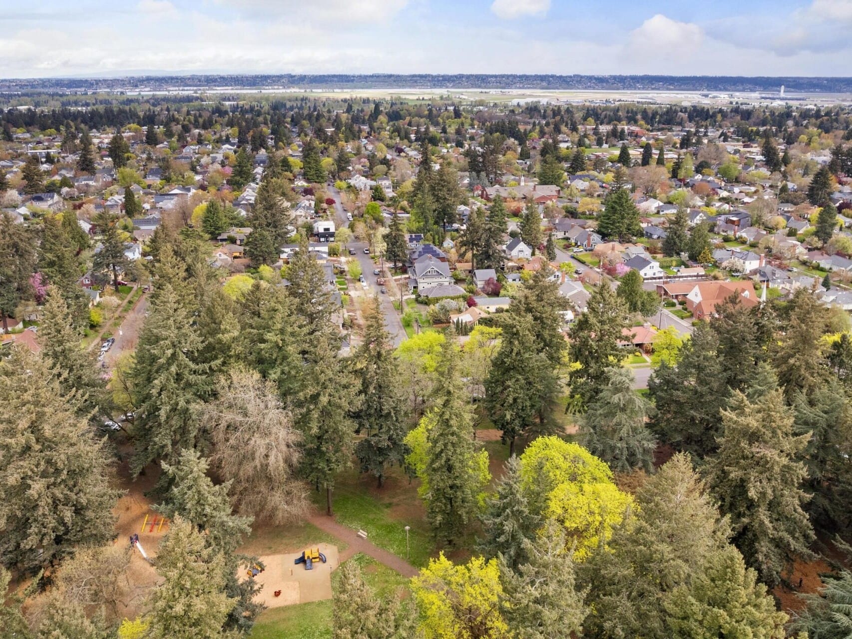 Aerial view of a suburban neighborhood boasting a mix of houses and lush greenery in Portland, Oregon. A park with tall trees and a playground graces the foreground, while rows of homes extend into the distance under a partly cloudy sky—a prime spot for any Portland Realtor to showcase.