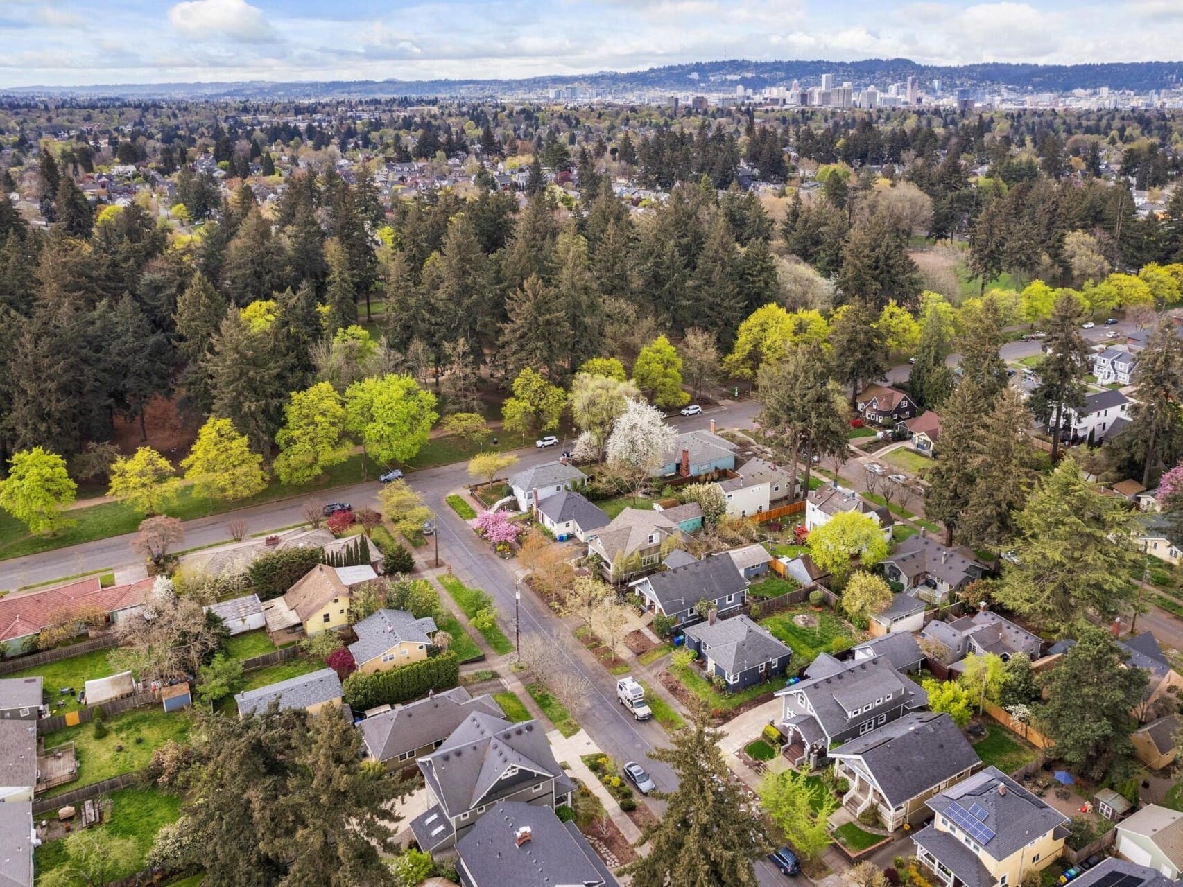 Aerial view of a residential neighborhood with tree-lined streets and houses in Portland, Oregon. A large park with dense trees graces the background, while the city skyline is visible in the distance under a partly cloudy sky, offering a glimpse of prime Portland real estate.