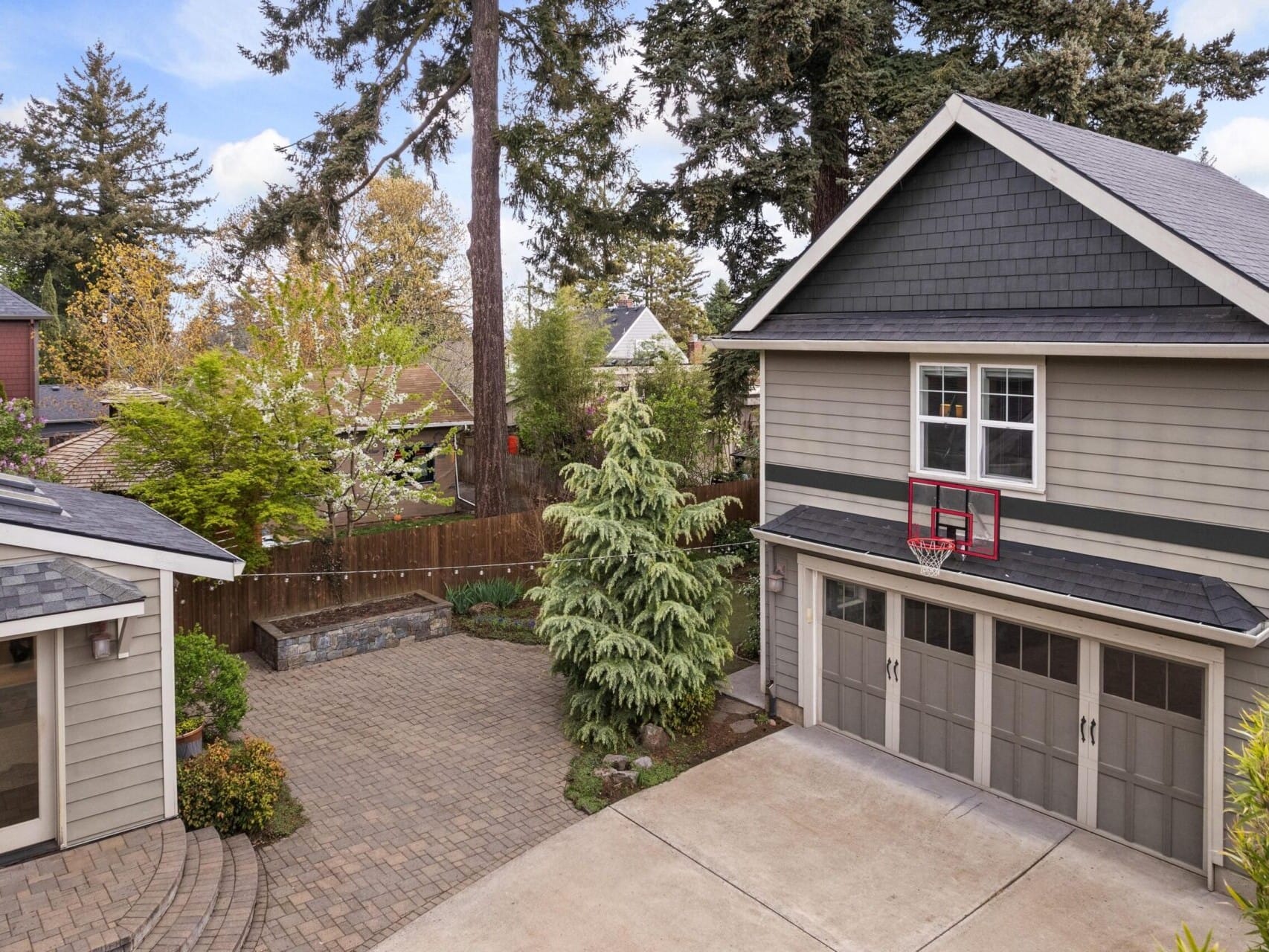 A paved driveway and yard with two gray buildings, featuring a garage with a basketball hoop above it. Tall trees and greenery surround the area under a clear blue sky—a picturesque gem in Portland Oregon Real Estate.