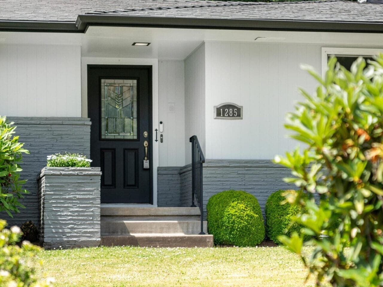 A modern house exterior with a gray and white color scheme. The entrance features a black door with glass panels and is flanked by greenery. The house number 1285 is displayed next to the door. Lush trees are visible in the background.