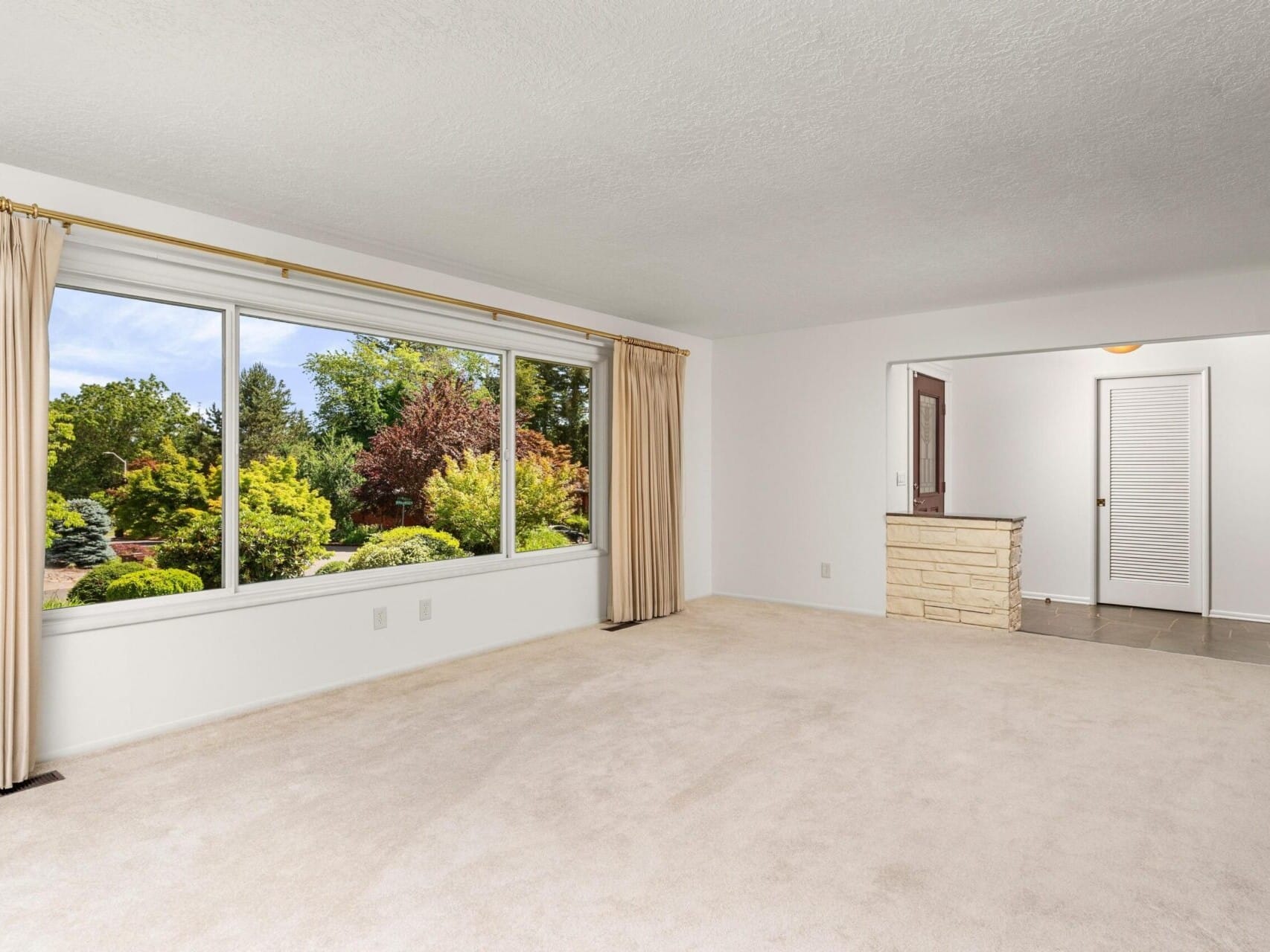 Bright and spacious empty room with beige carpet, large window with beige curtains overlooking a lush garden. An open doorway leads to a small area with a stone wall and a closed white door.