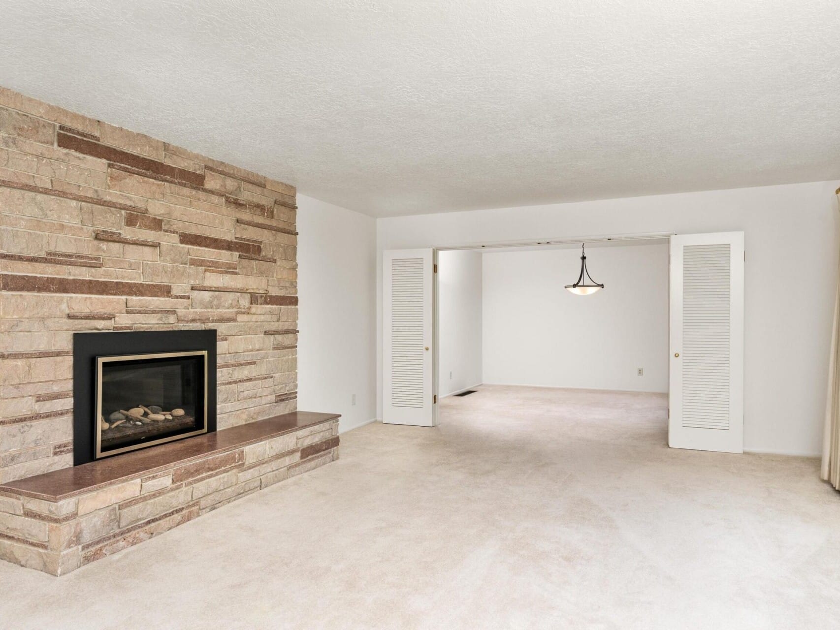 A spacious living room with beige carpeting features a stone fireplace on the left wall. Open double doors lead to an adjacent room with a hanging light fixture. Cream-colored walls and curtains frame the space.