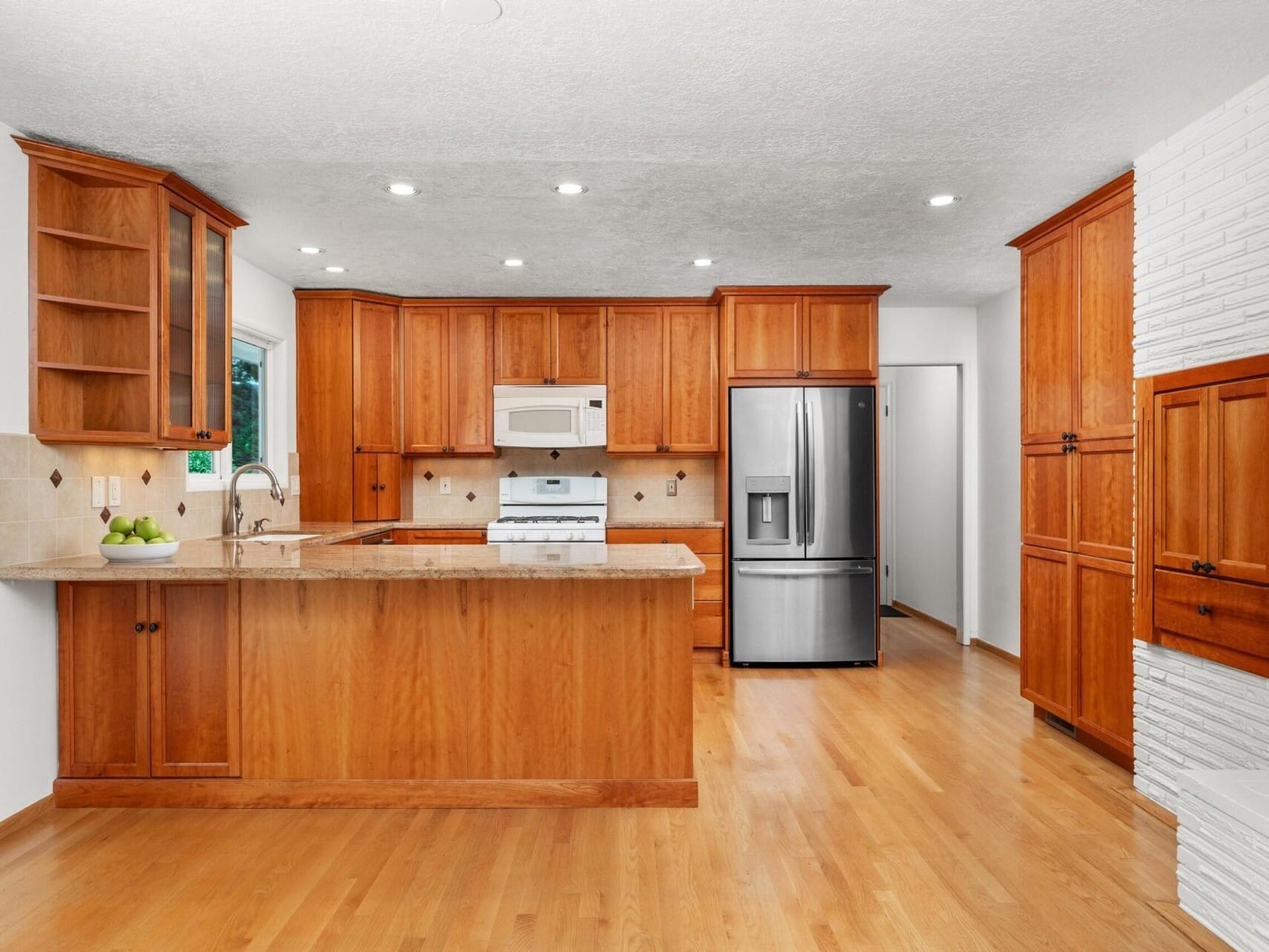 A spacious kitchen with wooden cabinets and a light wood floor. It features a stainless steel refrigerator, a white microwave and oven, and a central island with a beige countertop. Recessed lighting brightens the room.