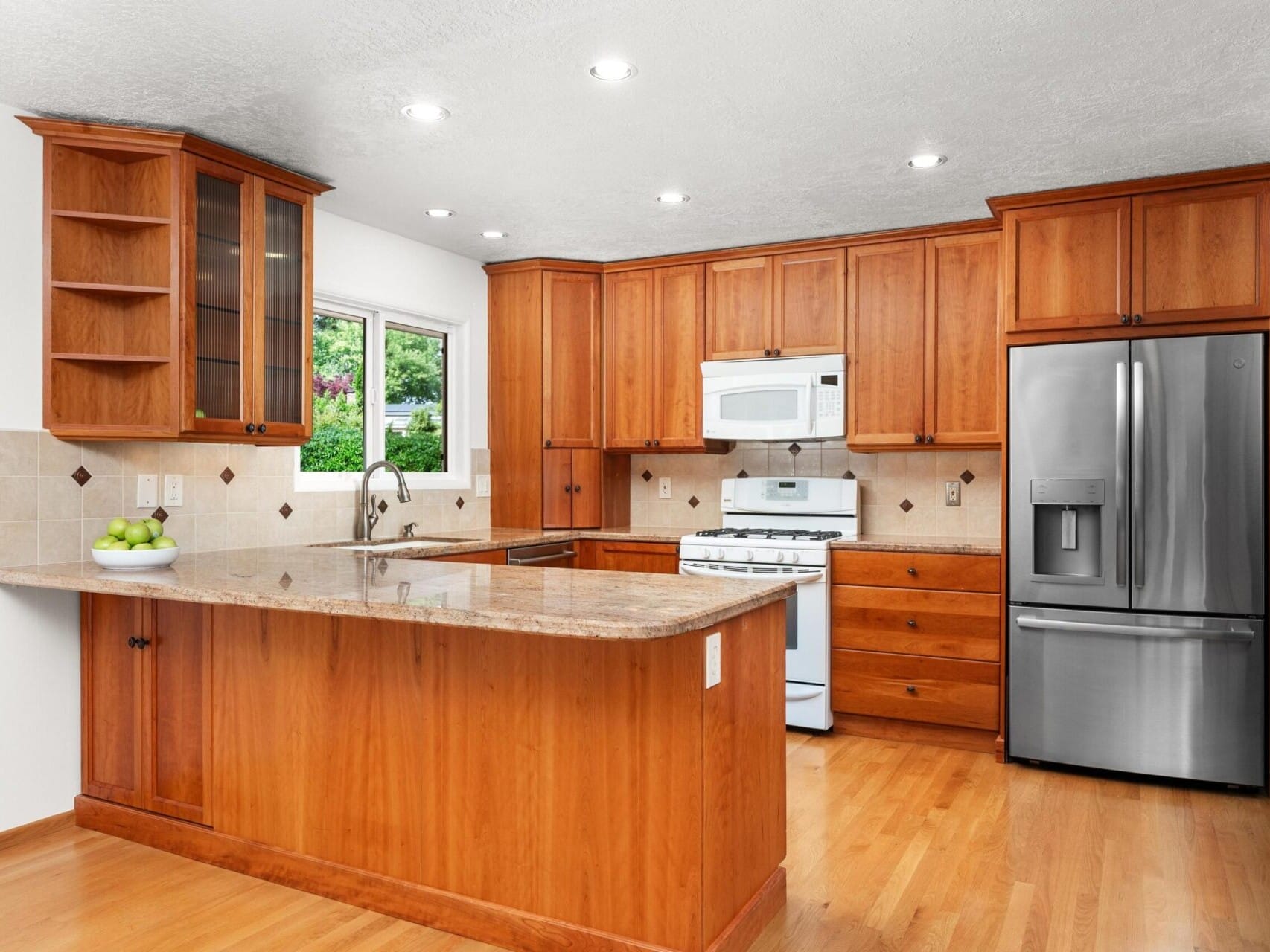 A spacious kitchen with wooden cabinets, a granite countertop island, and hardwood floors. It features a stainless steel refrigerator, a white microwave, and an oven. A window above the sink lets in natural light.