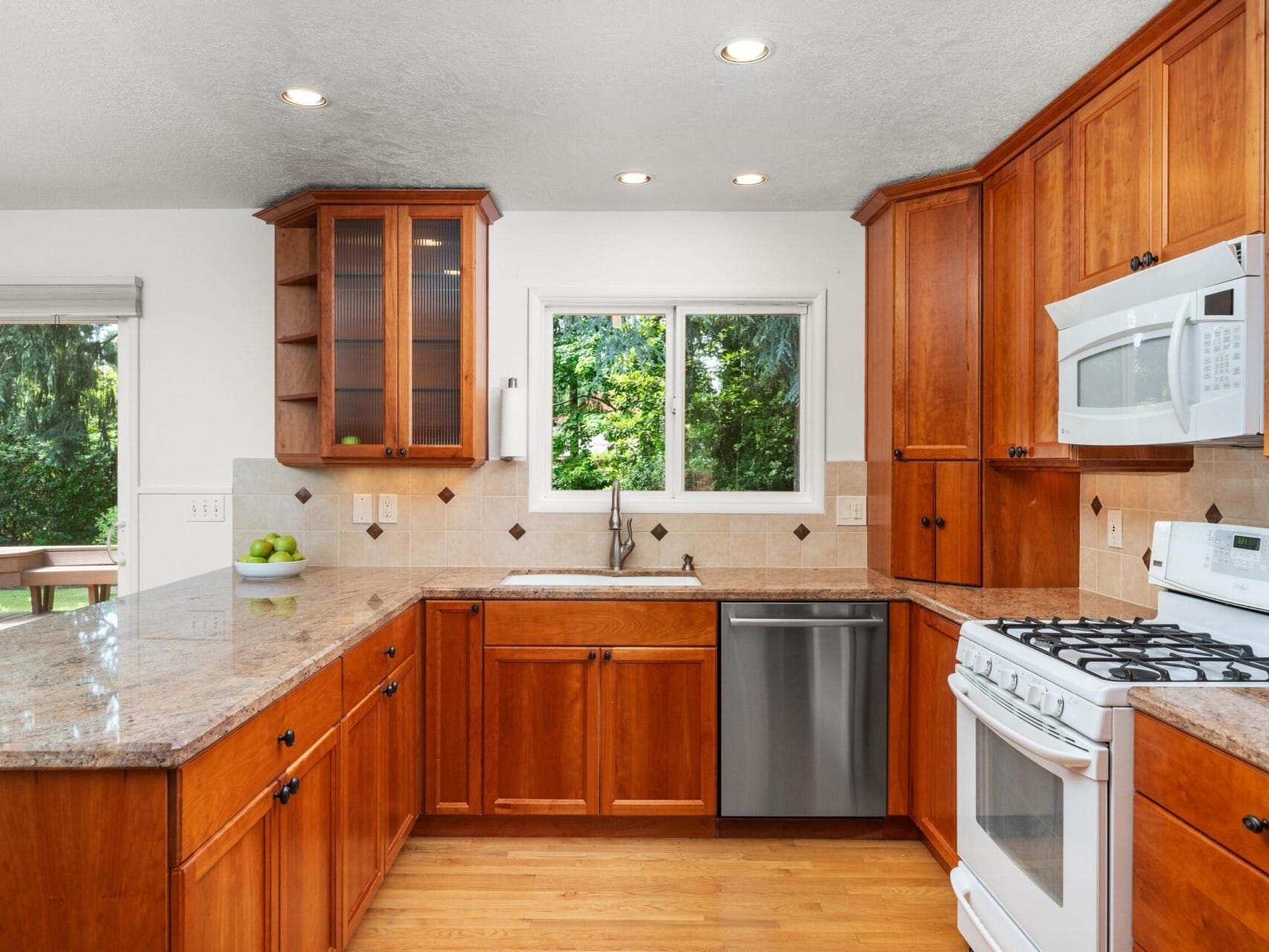 A kitchen with wooden cabinets, a granite countertop, and stainless steel appliances including a dishwasher and gas stove. A window over the sink offers a view of green trees outside. A bowl of green apples sits on the counter.