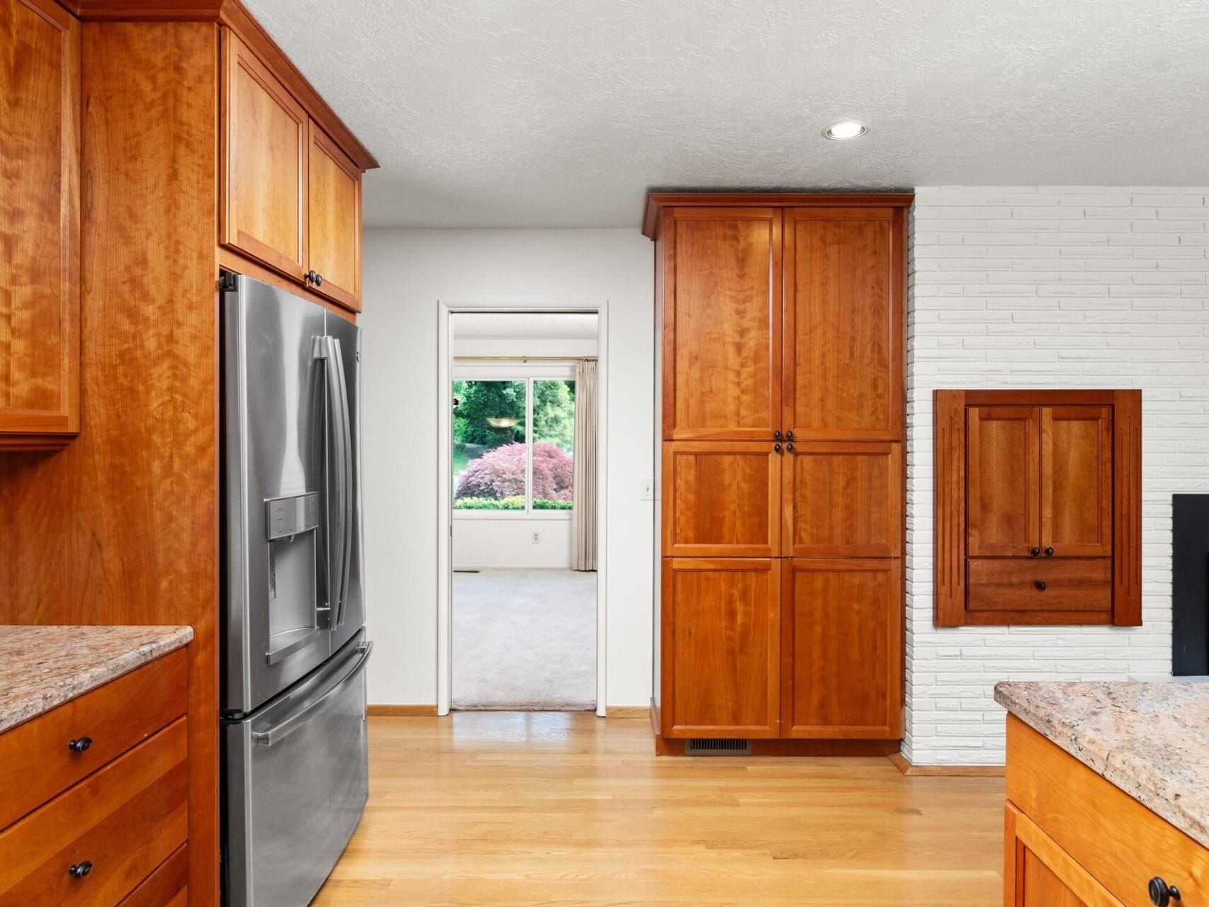 Modern kitchen with wooden cabinets, stainless steel refrigerator, and granite countertops. A doorway leads to a room with a large window overlooking a garden. Light wood flooring and white brick wall accent the space.