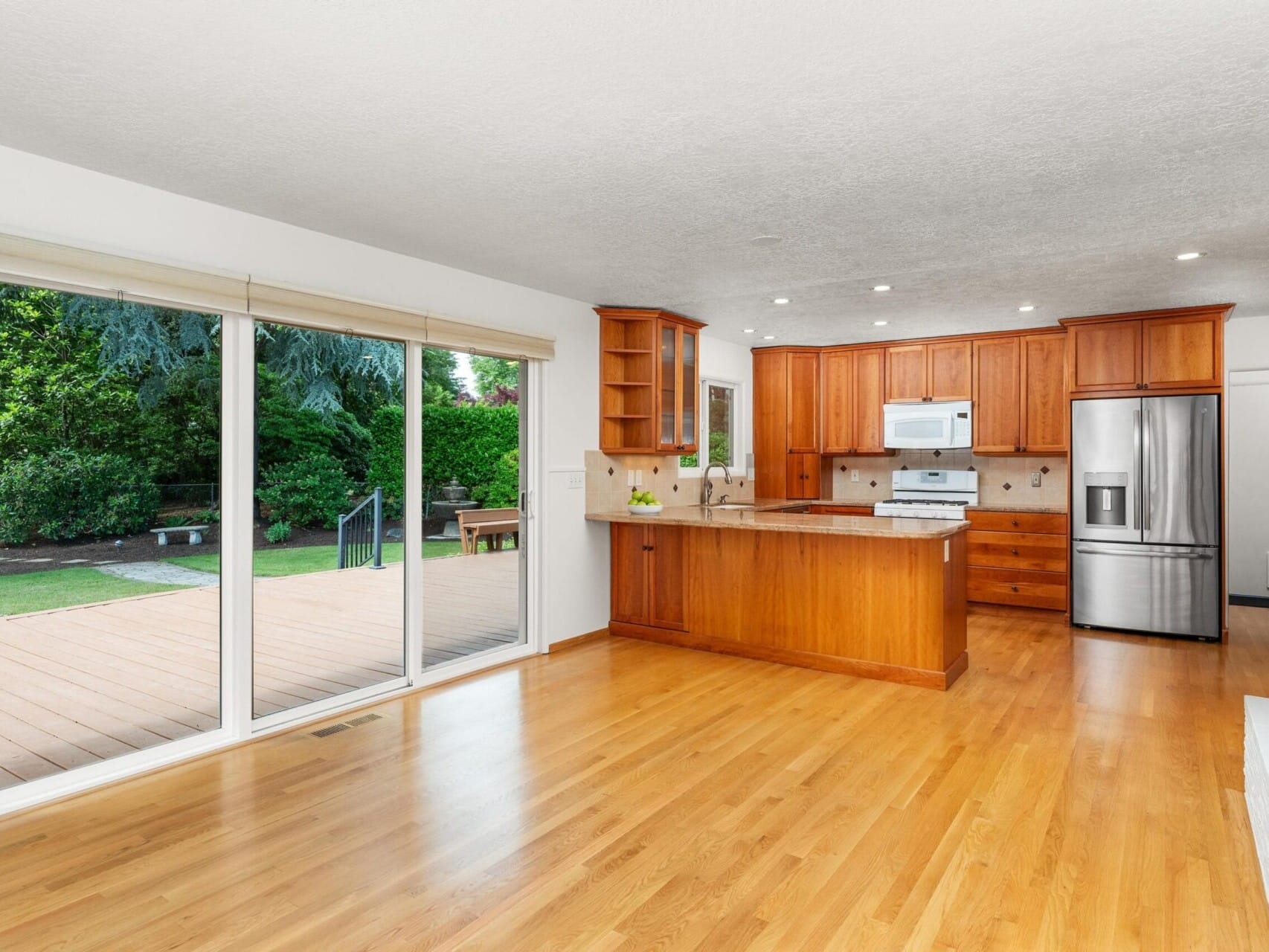 A spacious kitchen with wooden cabinets and a white countertop island. Stainless steel appliances are visible. Large windows and a sliding glass door open to a deck, providing ample natural light and a view of a garden with trees and greenery.