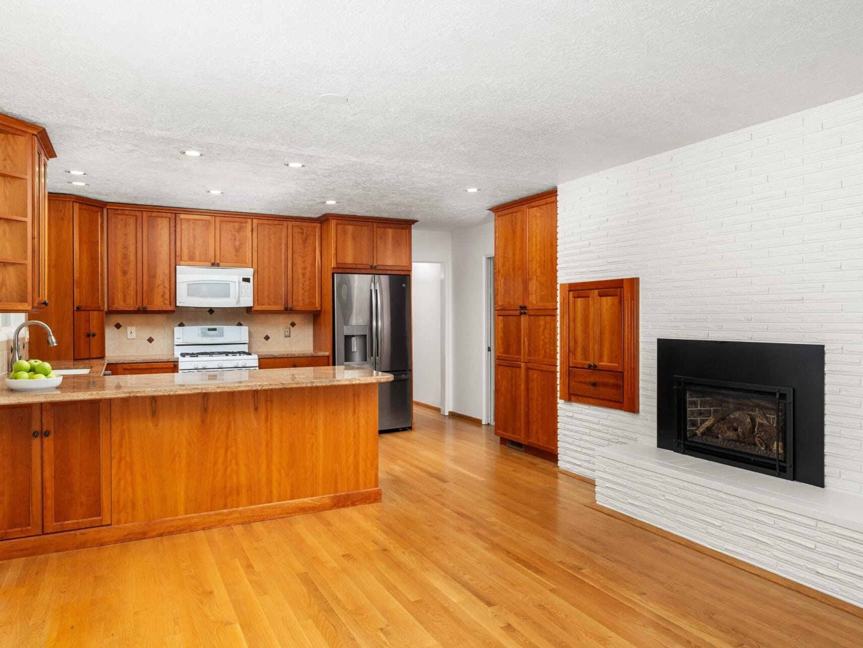 A modern kitchen with wooden cabinetry, stainless steel appliances, and a kitchen island. Adjacent is a white brick wall with a built-in fireplace. The floor is light wood, contributing to a warm, inviting atmosphere.