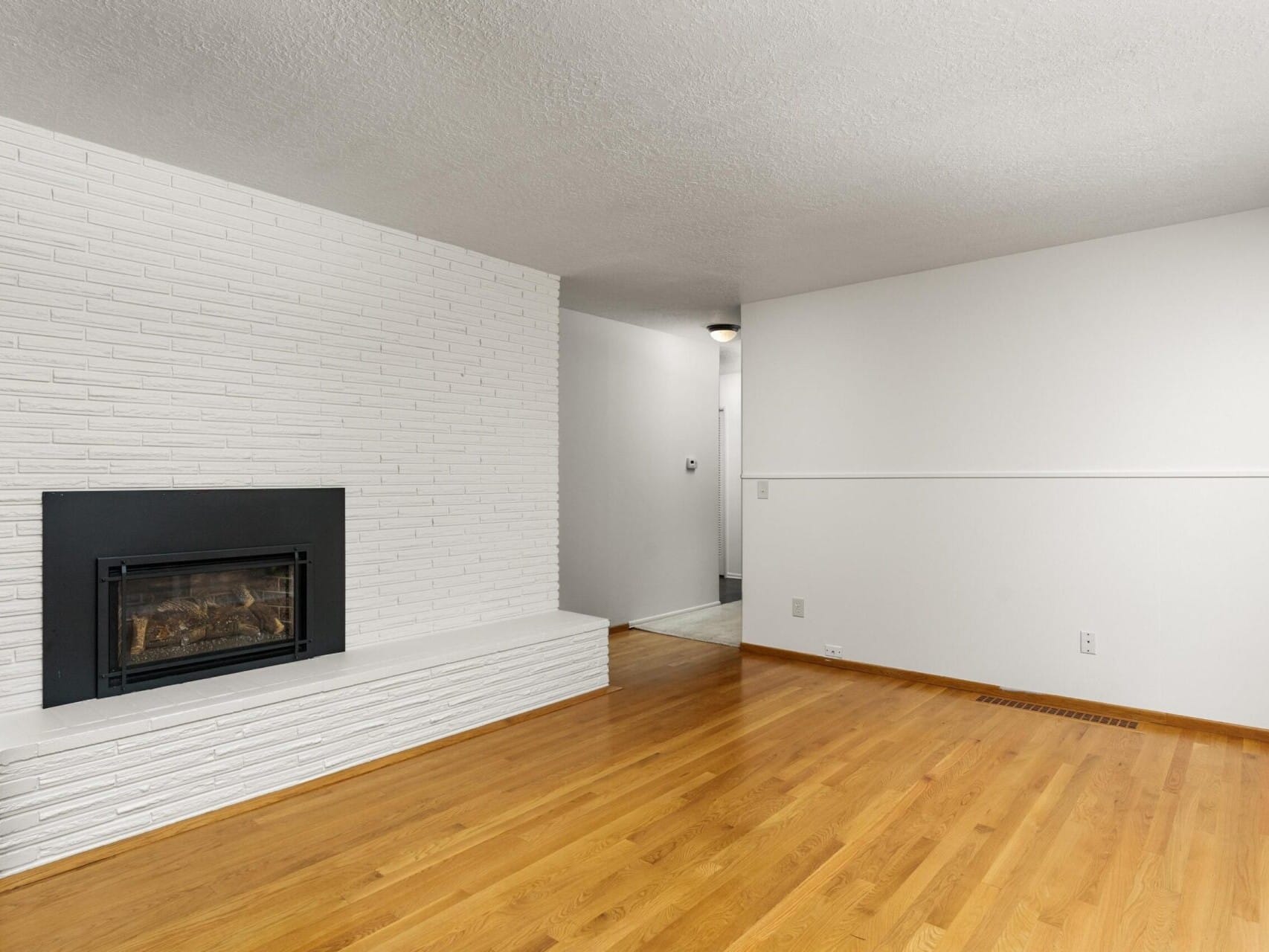 A minimalist living room with a light wood floor and white brick fireplace. The room features a textured white wall, a small recessed light fixture in the hallway, and an empty wooden mantel beside the fireplace.