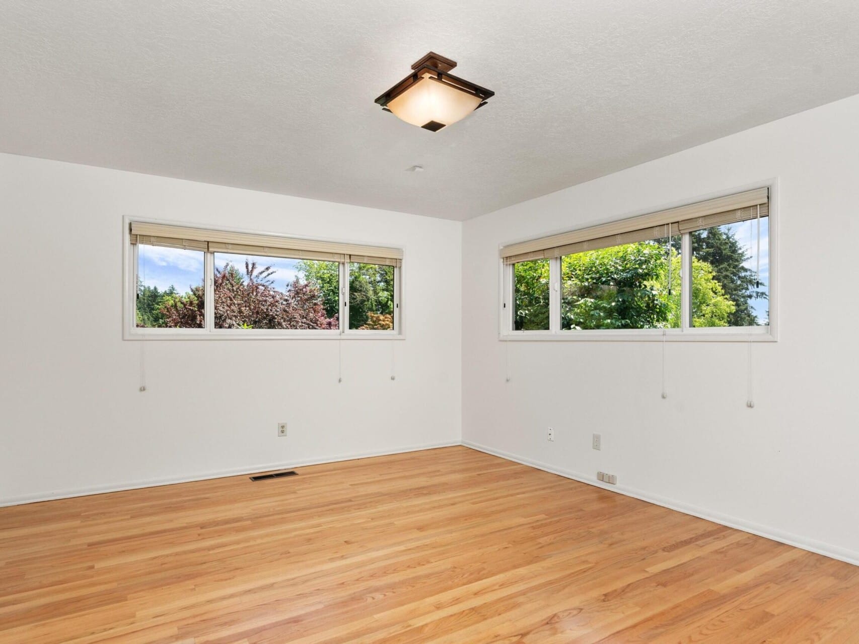 An empty room with white walls and hardwood flooring. It features two large windows with beige blinds, offering a view of trees and a clear sky outside. A square ceiling light fixture is centered on the ceiling.