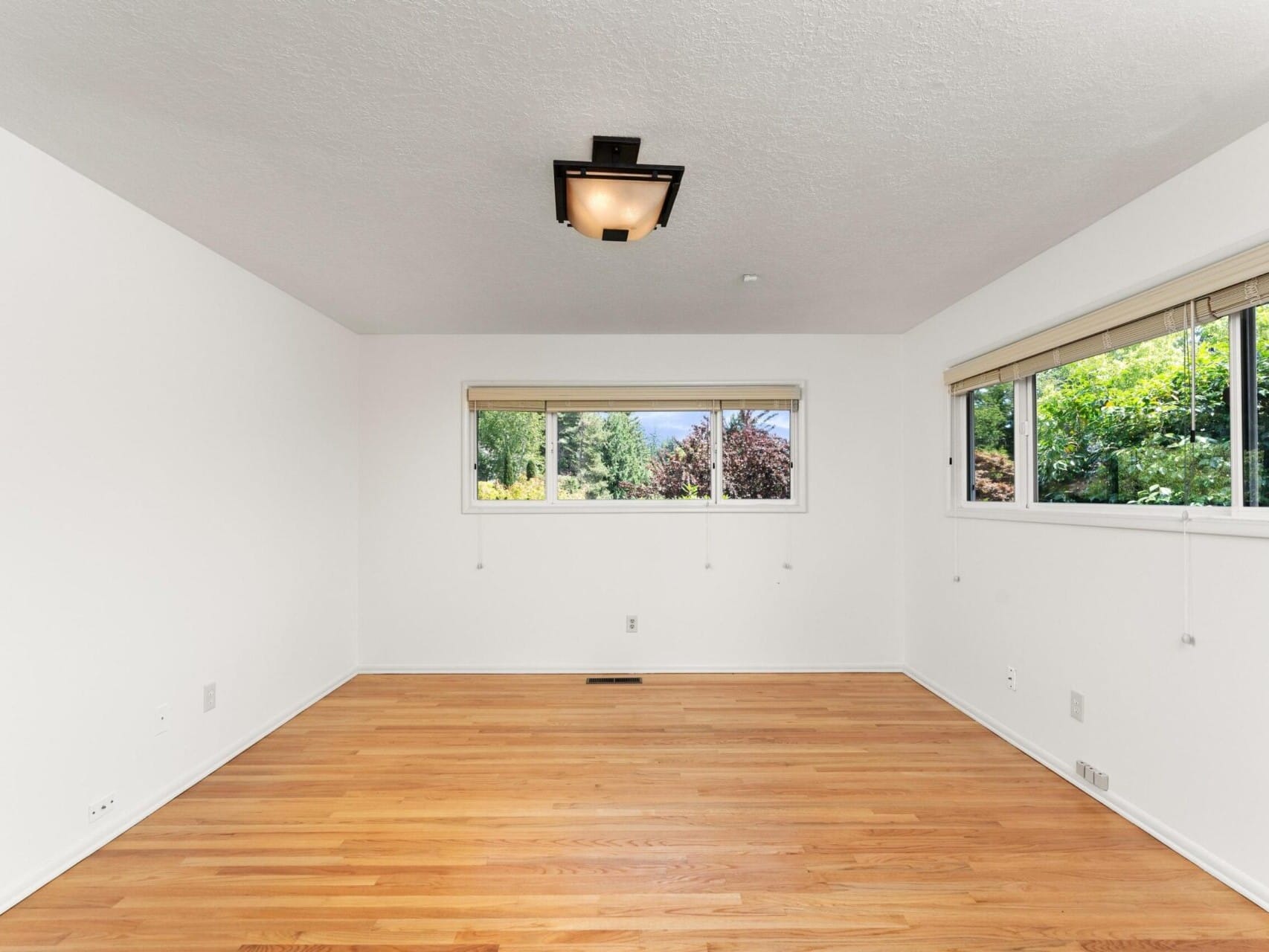 A bright, empty room with white walls, hardwood flooring, and a rectangular ceiling light. Three large windows provide a view of lush greenery outside, allowing natural light to fill the space.