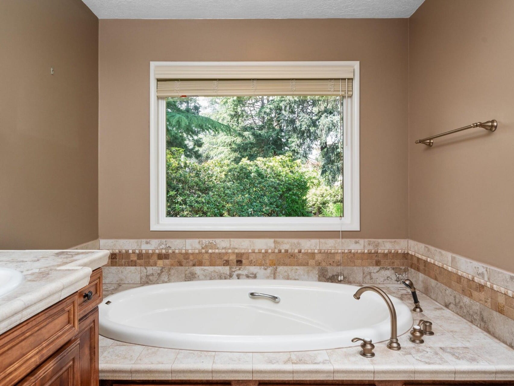 A bathroom with a large white oval bathtub surrounded by beige and brown tiles. A wooden cabinet is visible on the left. A window above the tub offers a view of lush green trees outside, and a towel rack is mounted on the right wall.