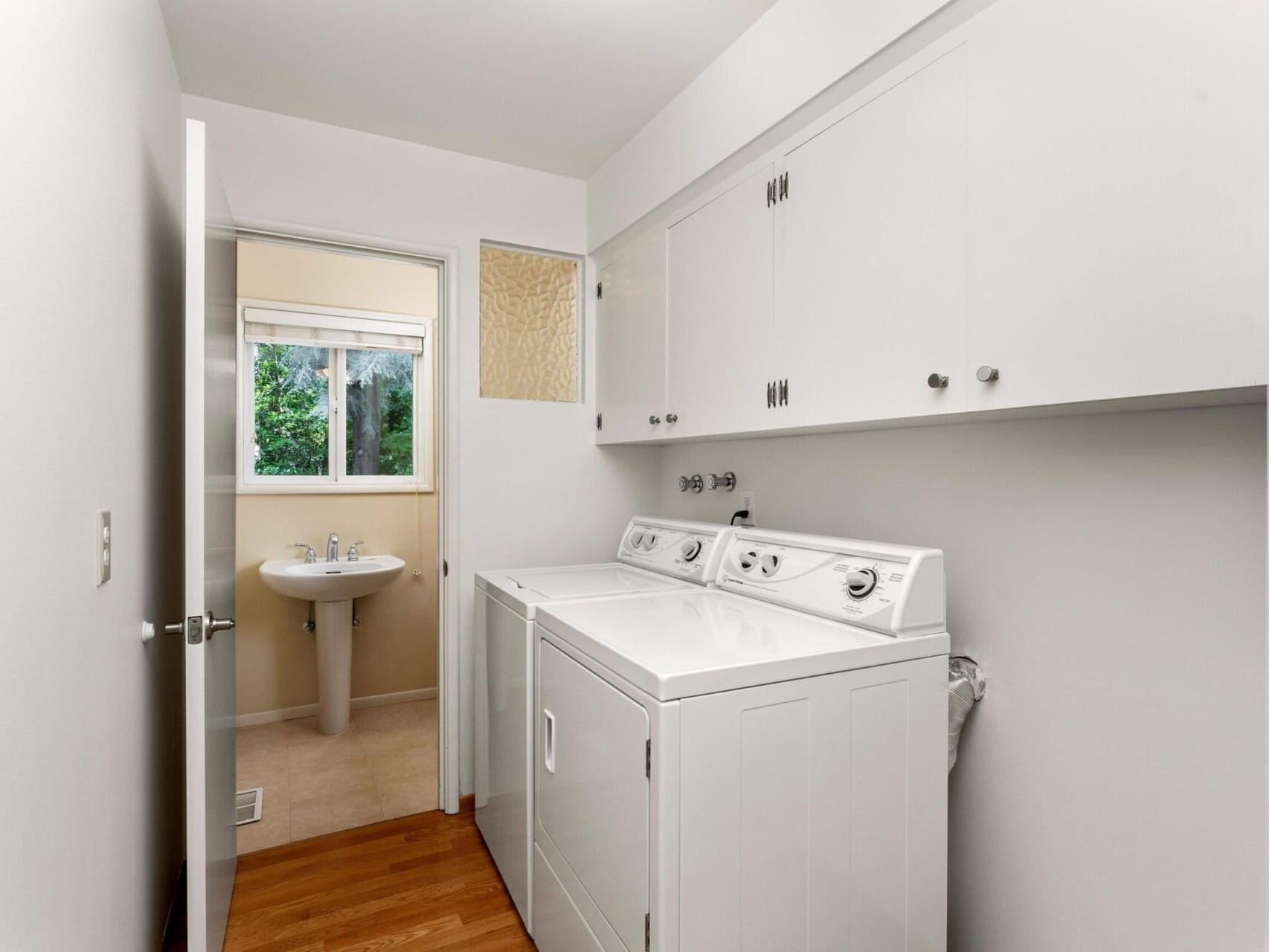 A compact laundry room with a white washing machine and dryer on the right. Cabinets are above the appliances. A small window with a view of greenery is in the background, and theres a pedestal sink near the window. Wood flooring is visible.