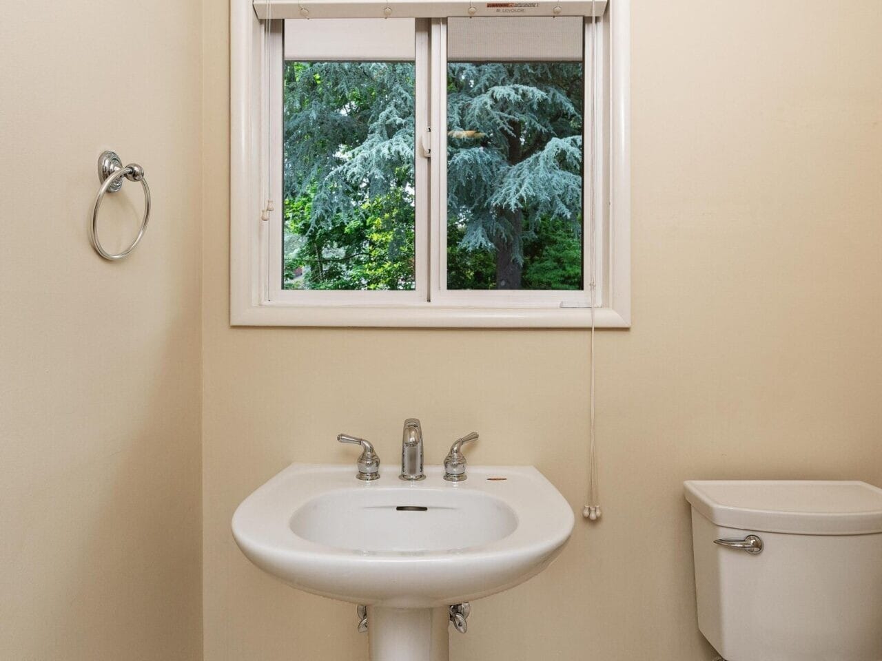 A small bathroom with a white pedestal sink and toilet. A window above the sink shows lush greenery outside. The walls are beige, and the floor is tiled. A towel ring is mounted on the left wall.